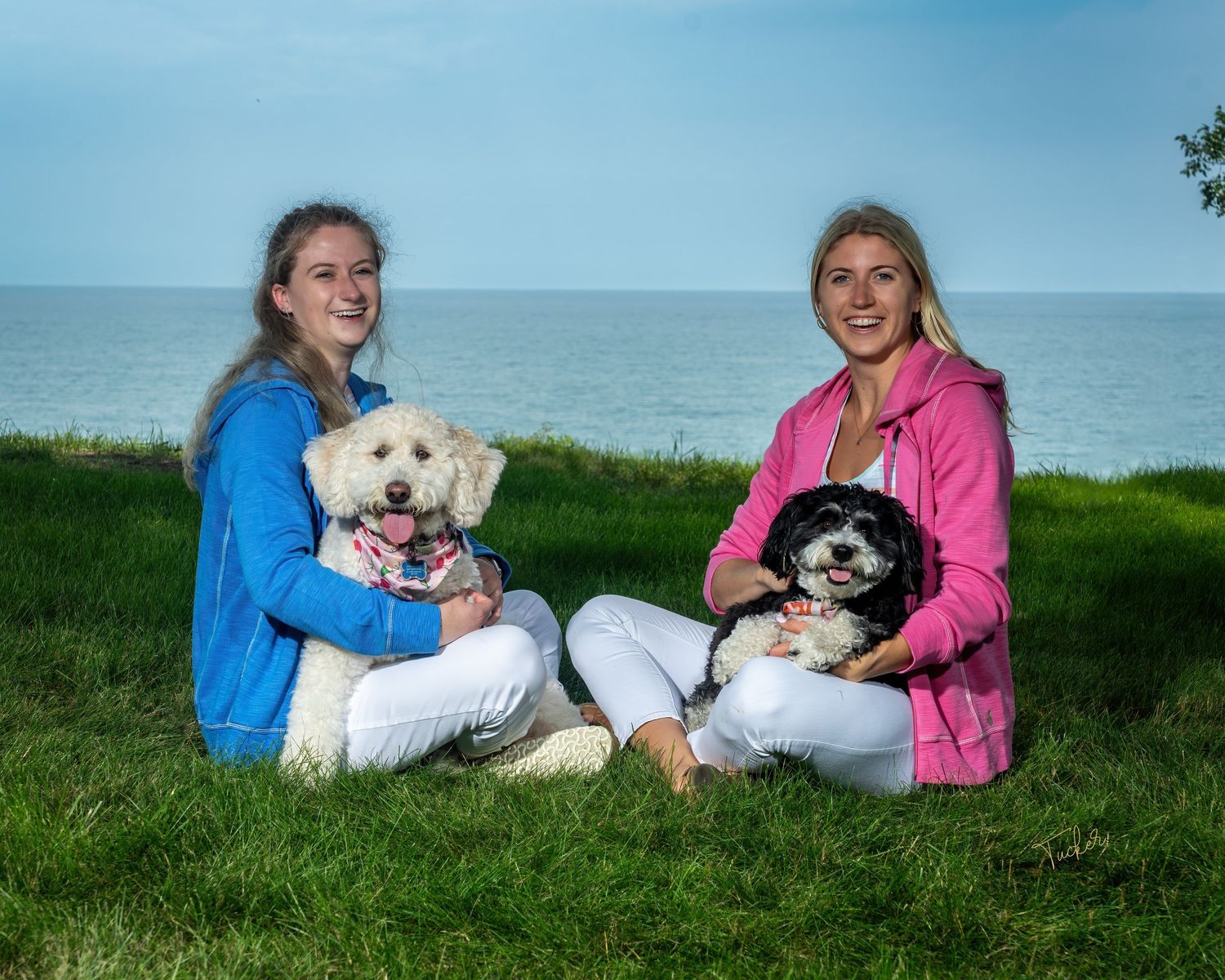 Two young women sitting on grass near lake, each holding a small fluffy dog in their laps.