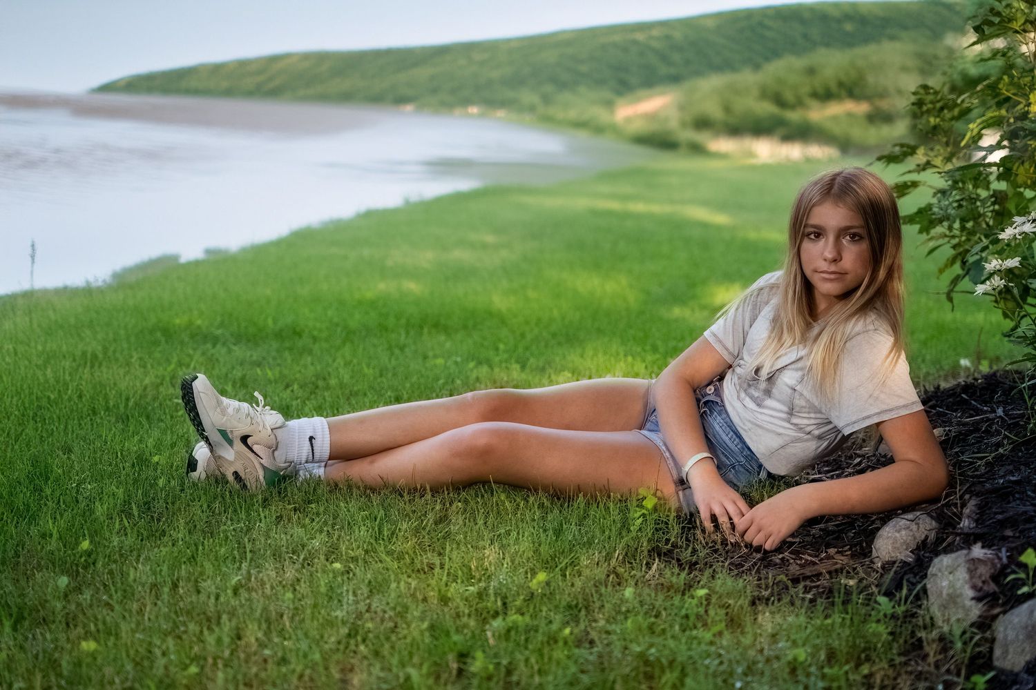 Teenage girl in denim shorts and white t-shirt relaxing on green grass by a calm lake at dusk.