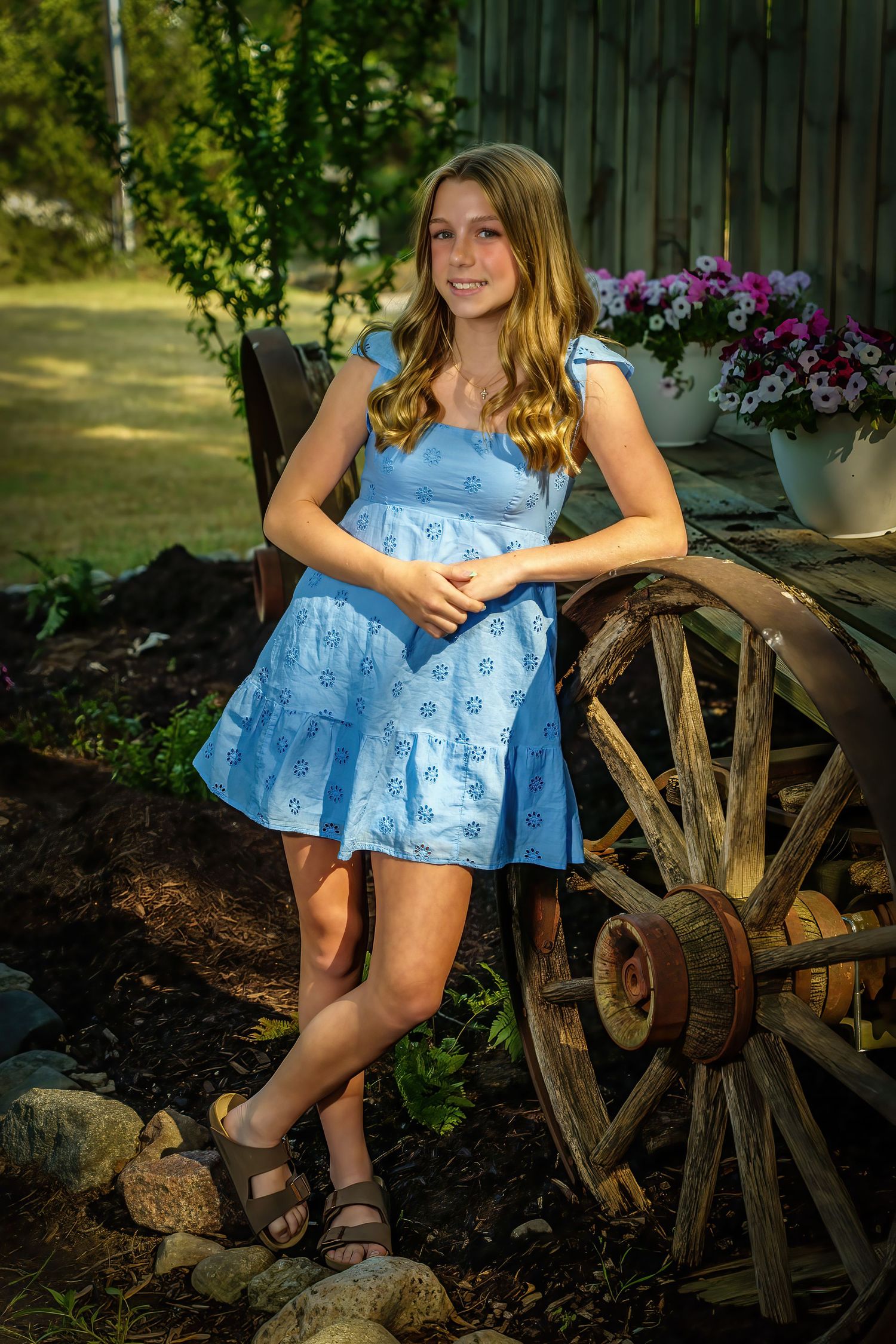 Teen girl in light blue dress leaning against rustic wooden wagon wheel surrounded by garden flowers.