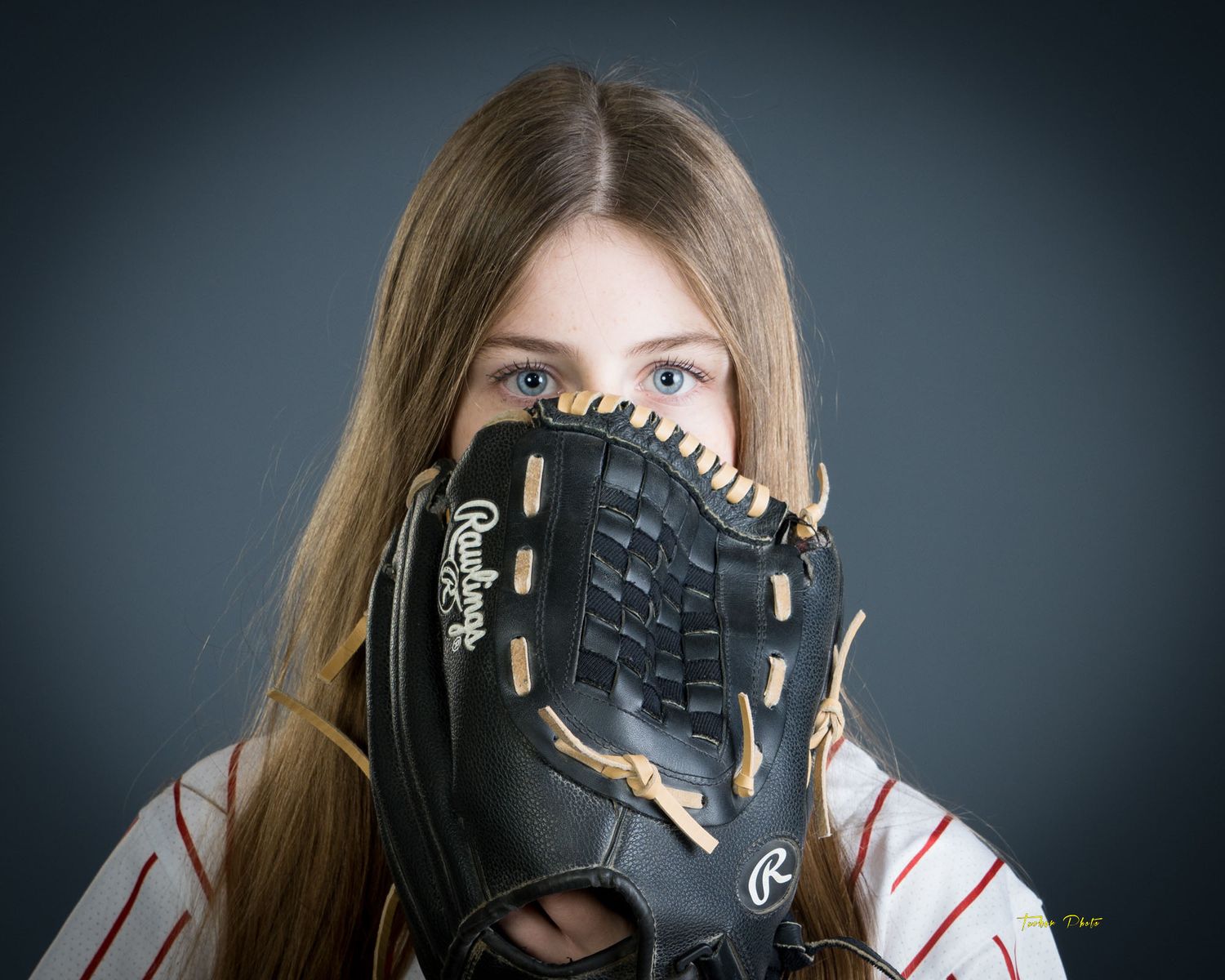 Young girl with long brown hair holding a Rawlings baseball glove up to her face, gray background.