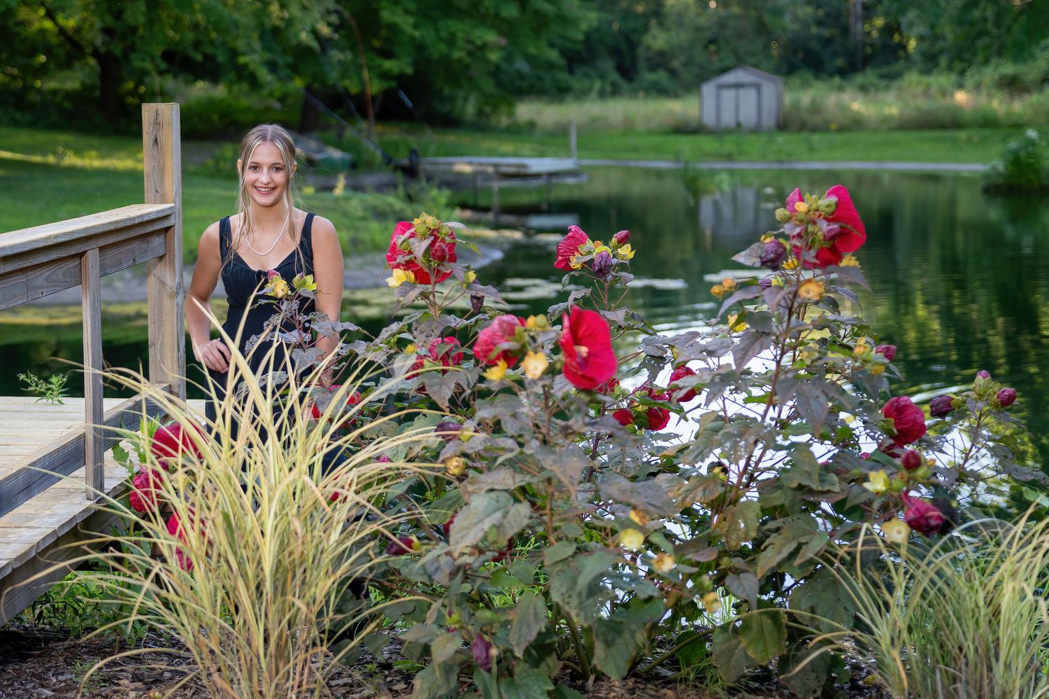 Smiling teen girl poses beside red flowers near a serene garden pond.