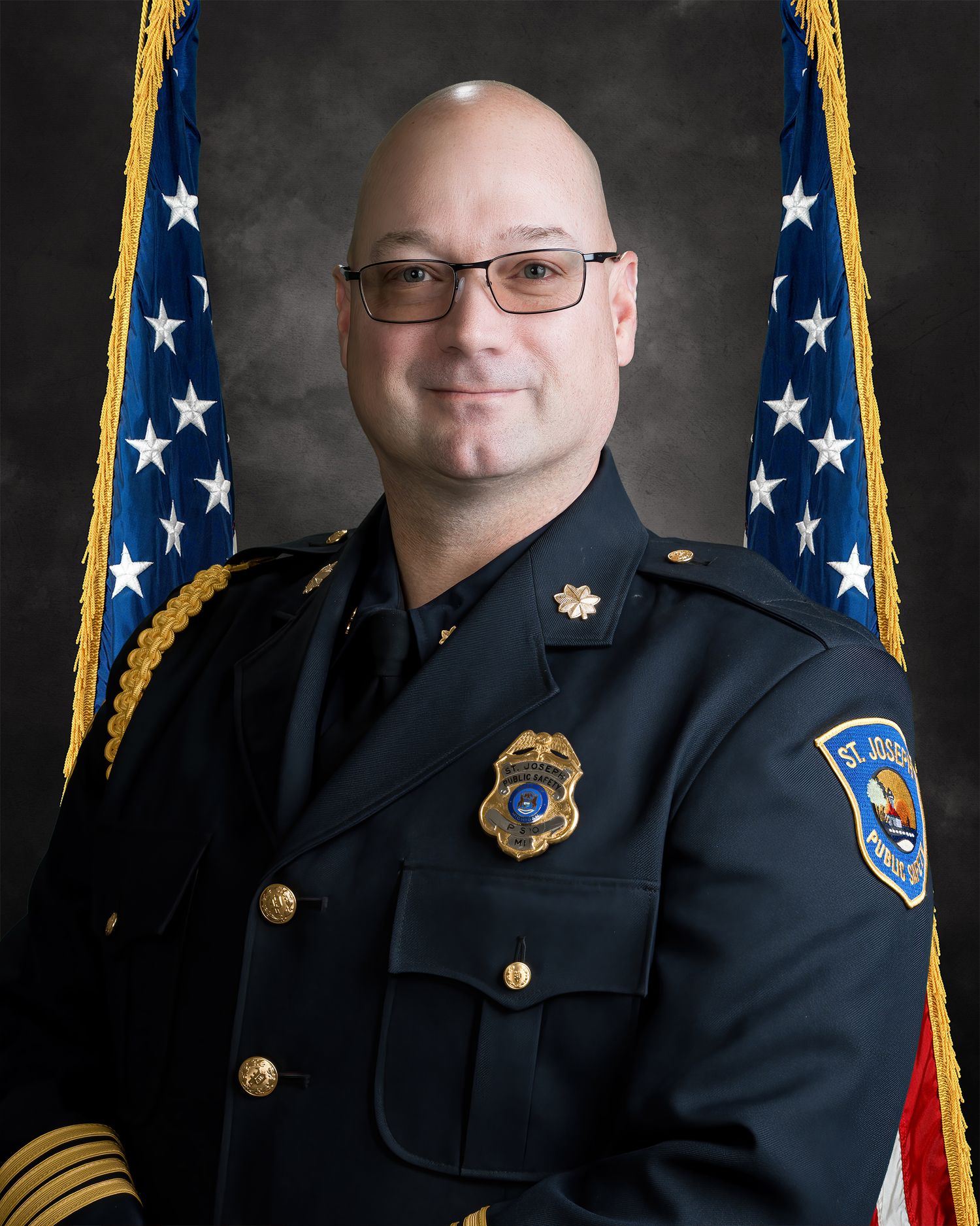 St. Joseph Public Safety officer in dress uniform with badge posing before American flags in official portrait.