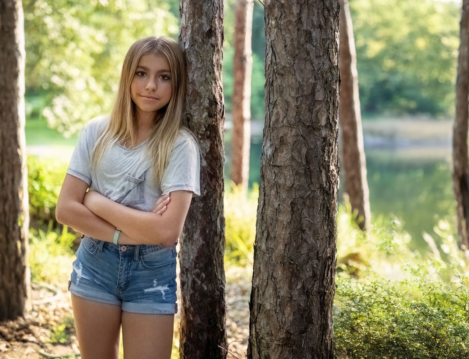 Teenage girl in denim shorts and white t-shirt standing between trees near a sunlit lake.