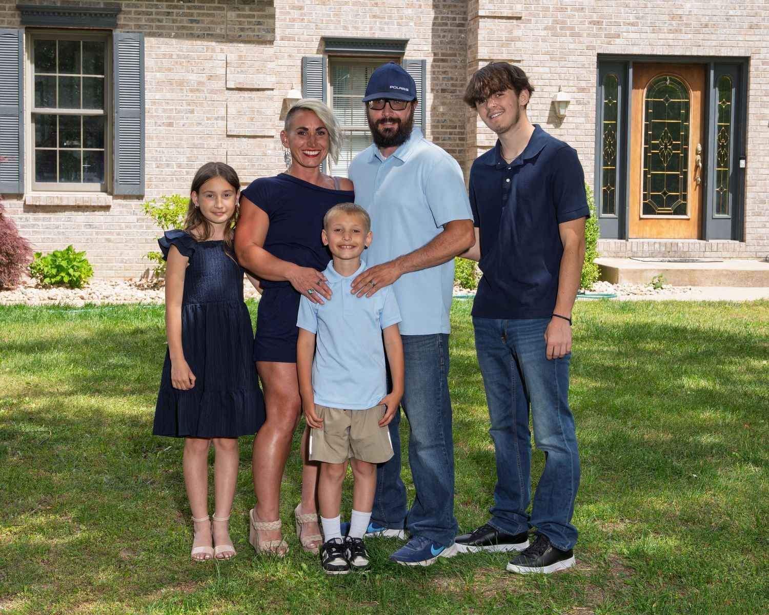 A family of five poses together on a green lawn in front of a large brick home on a sunny day.