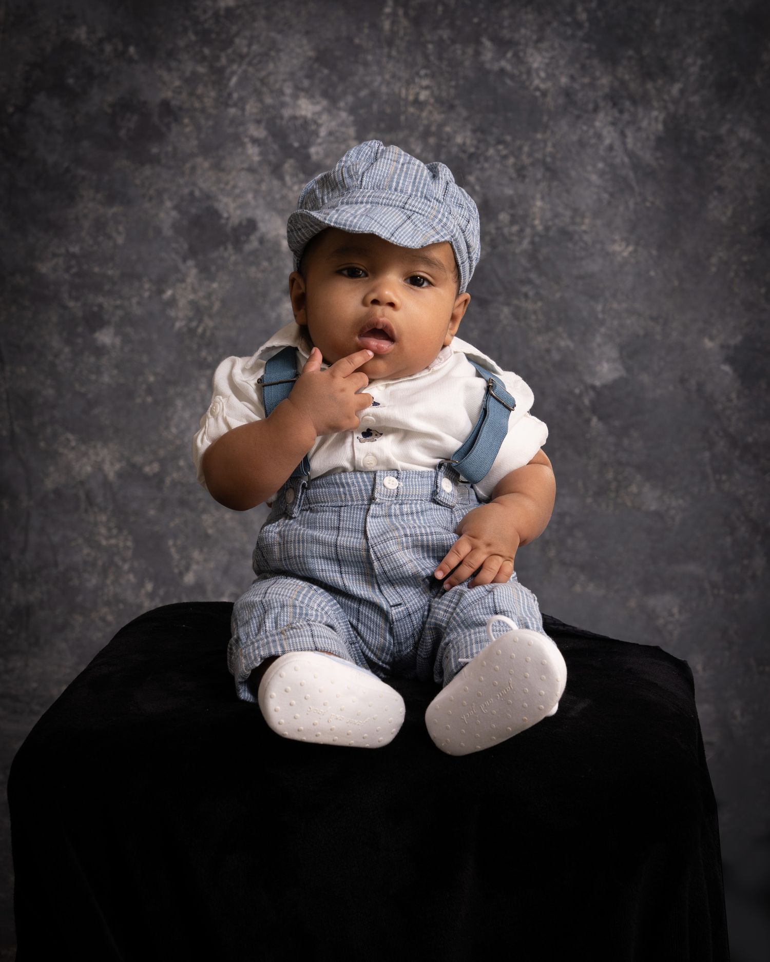 Adorable baby boy in blue plaid overalls and newsboy cap in studio portrait.