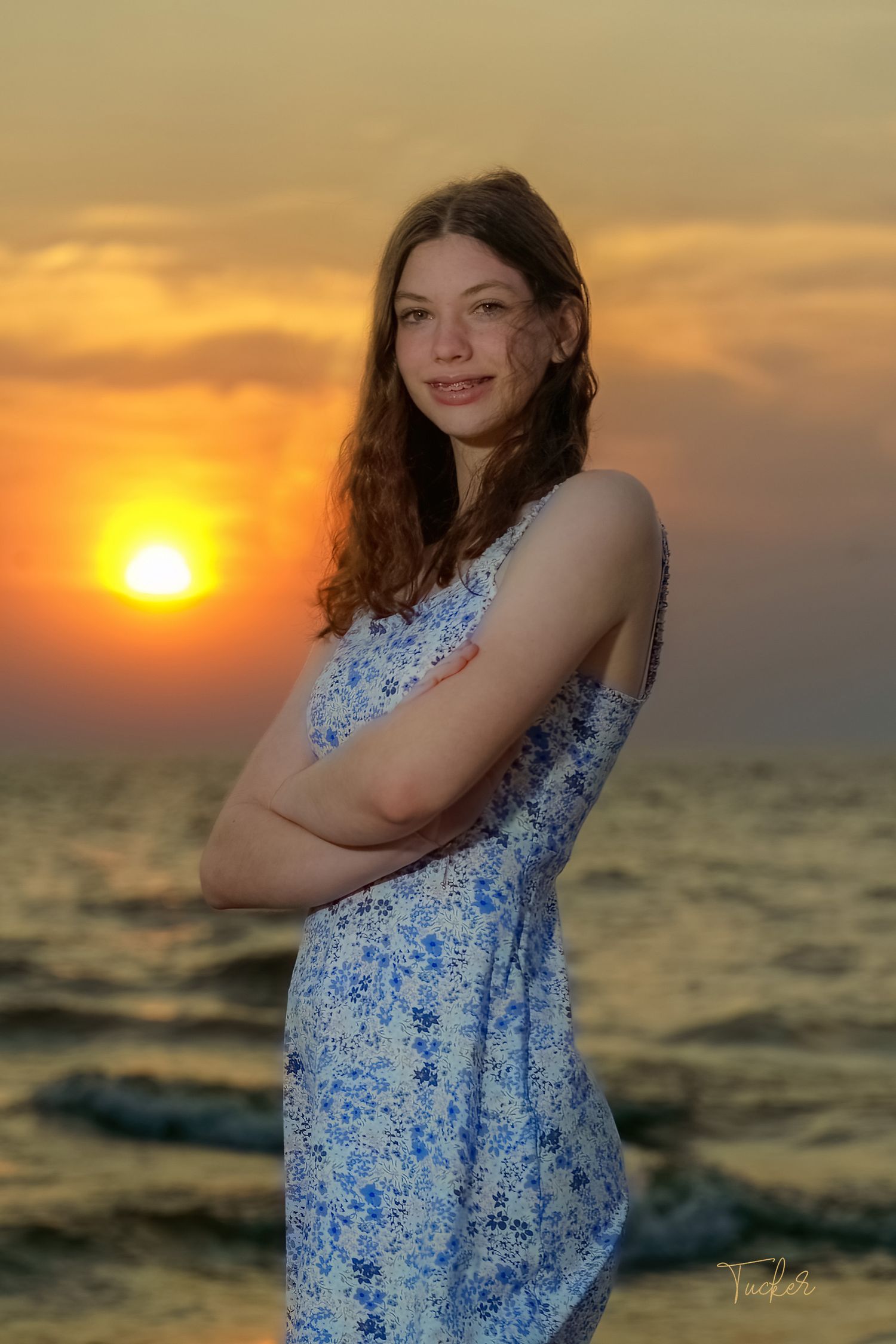 Smiling teenage girl in floral sundress standing on beach with vibrant orange sunset behind her.