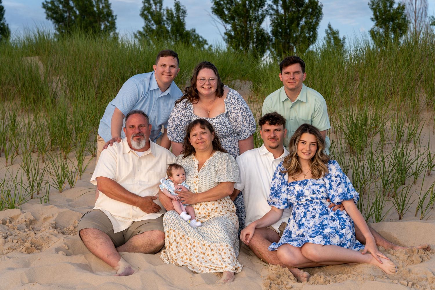 Extended family of seven posing together on sandy beach dunes with green grass in the background.
