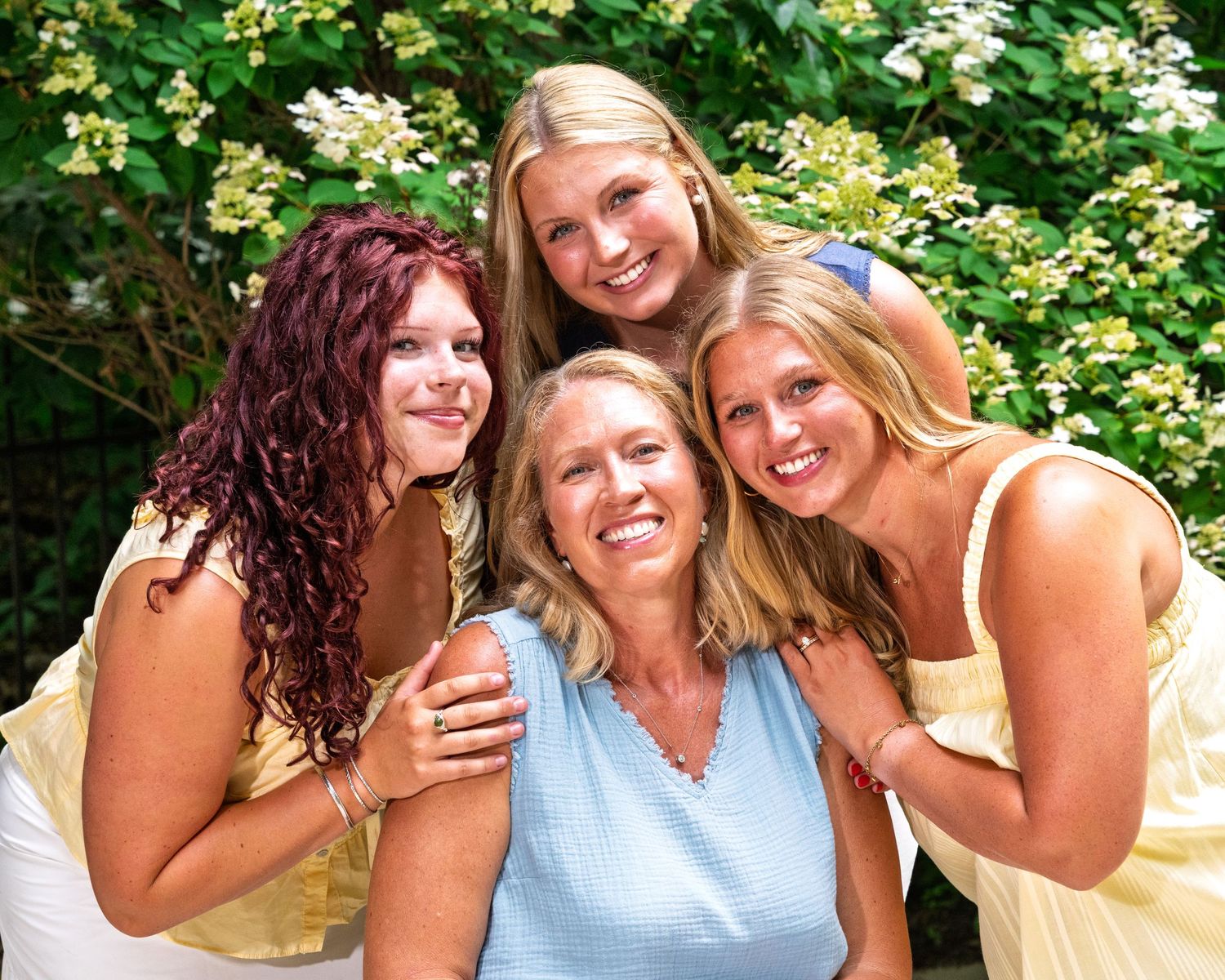 A smiling mother poses outdoors with her three blonde and brunette daughters surrounded by lush green foliage.