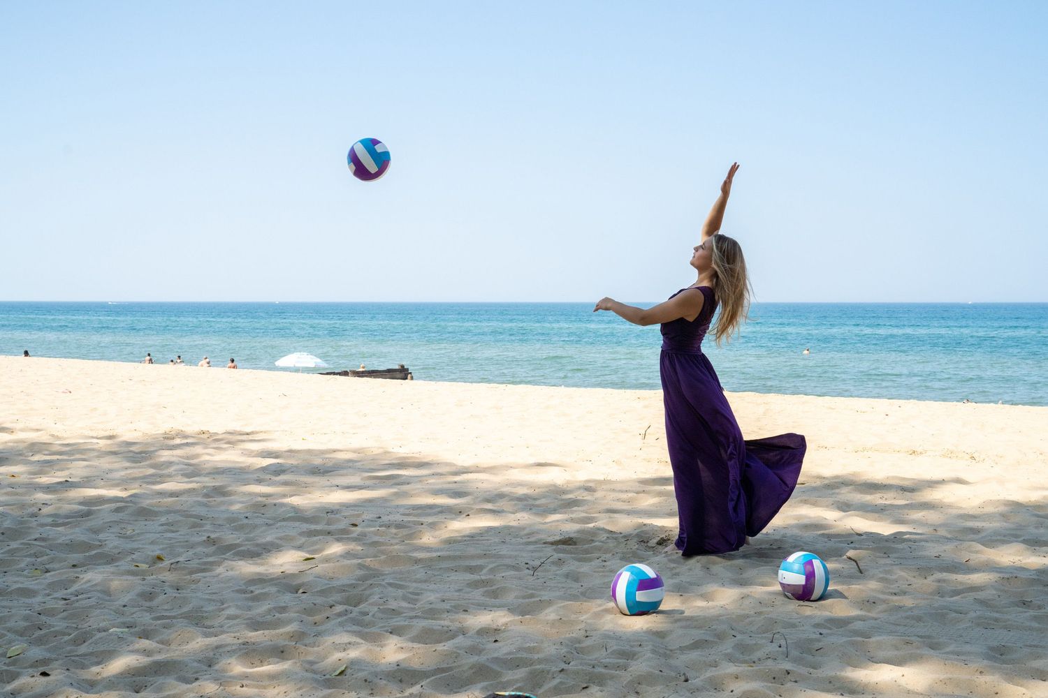 Woman in purple maxi dress tosses volleyball on sunny sandy beach with ocean in background.
