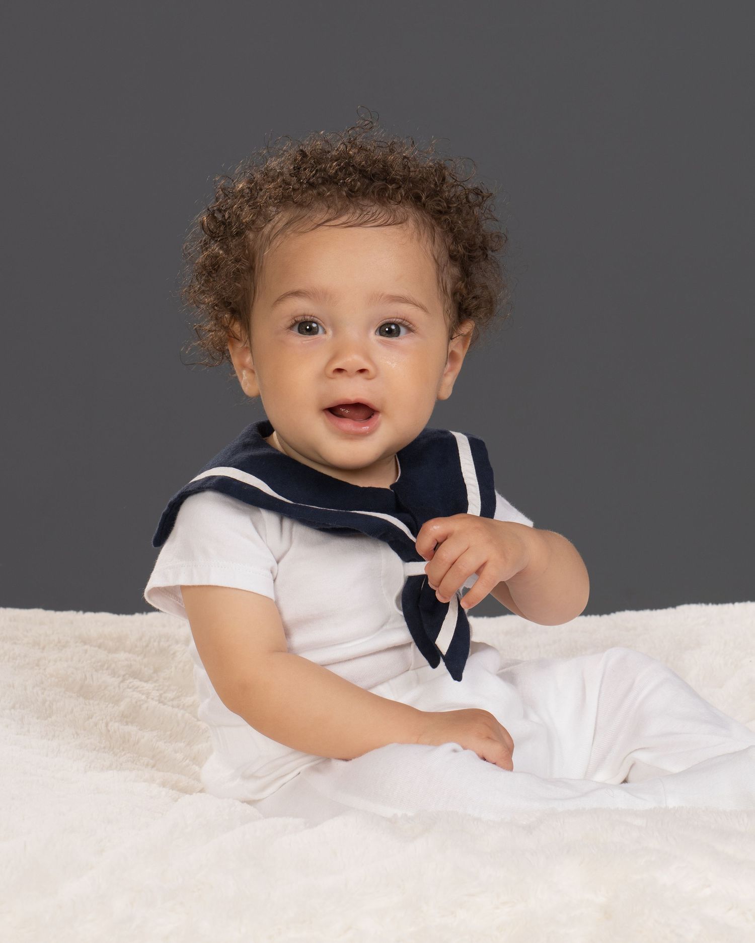 Happy curly-haired baby in sailor outfit smiling in professional studio portrait photography.