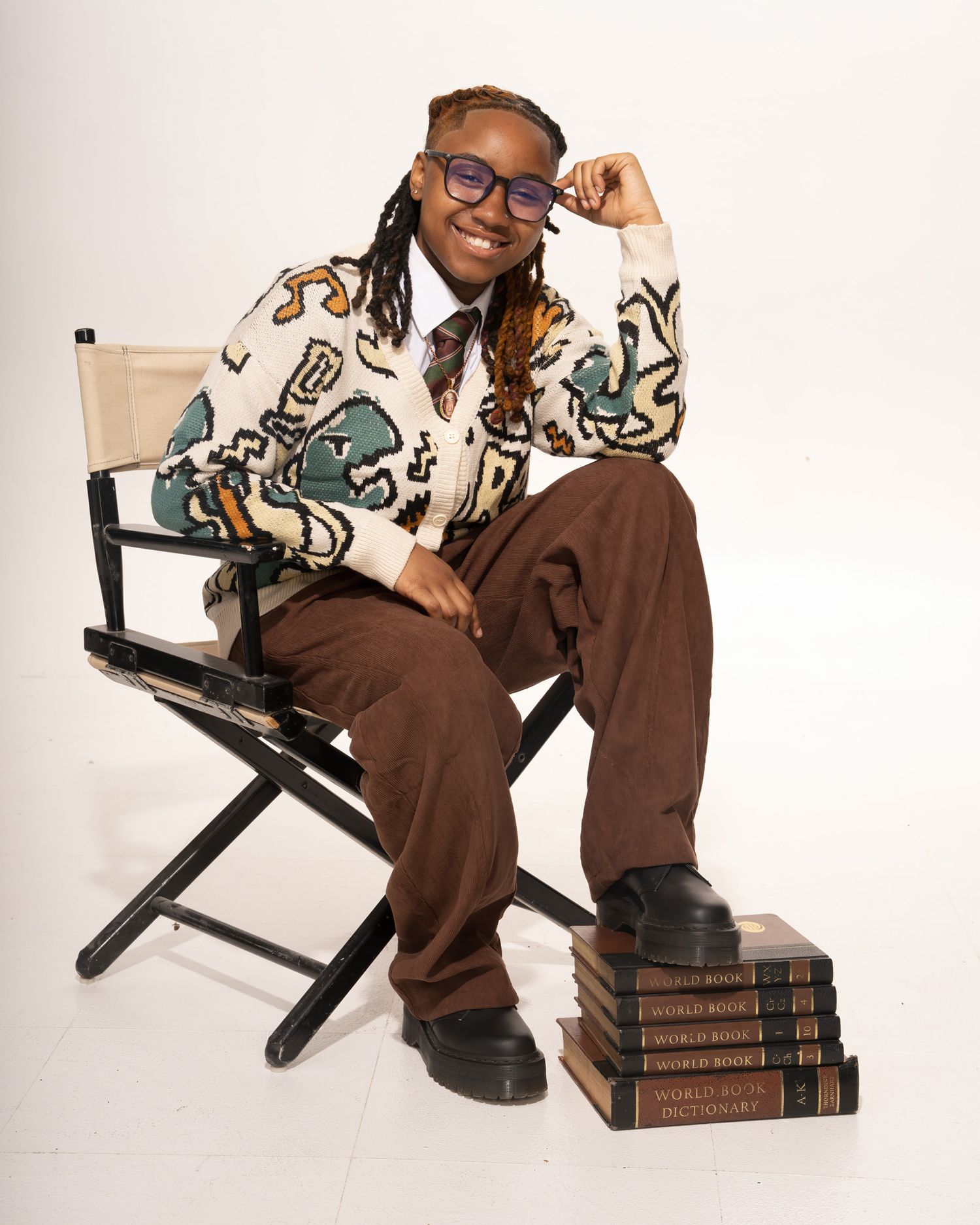Smiling student sitting in director chair with stacked World Book encyclopedias wearing graphic cardigan.