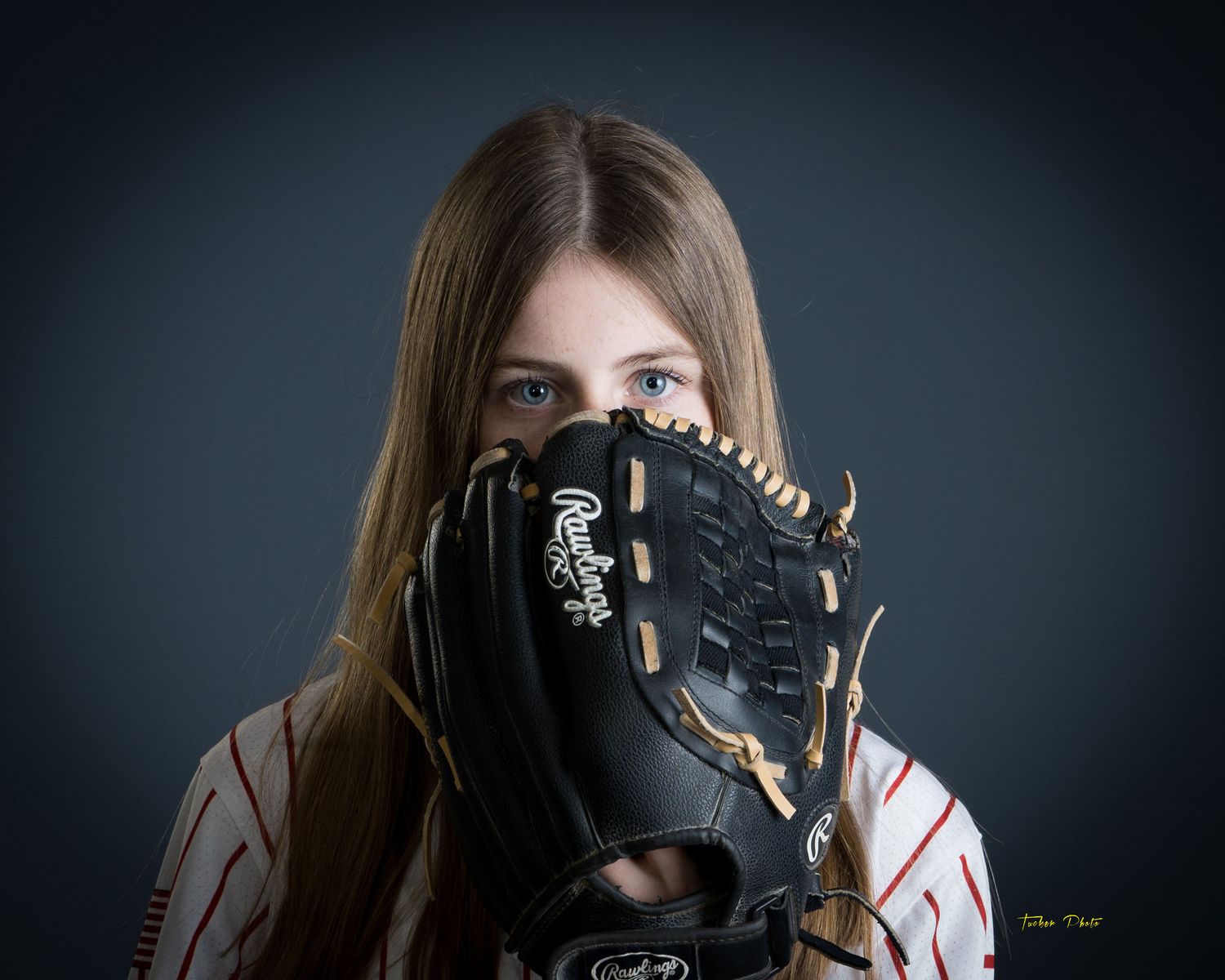 Young female baseball player with long hair holding a Rawlings mitt up to her face, gray background.