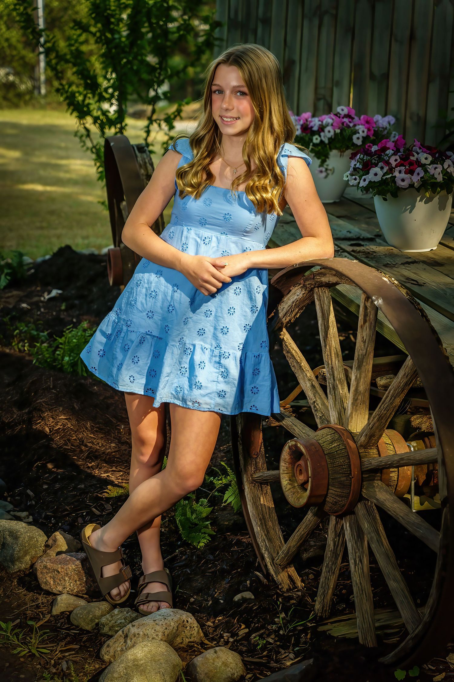Smiling teen in blue floral dress posing beside antique wooden wagon wheel with white potted flowers.