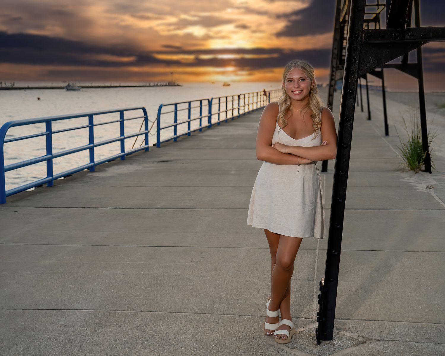 Smiling blonde teen in white dress leaning on pier post at stunning waterfront sunset.