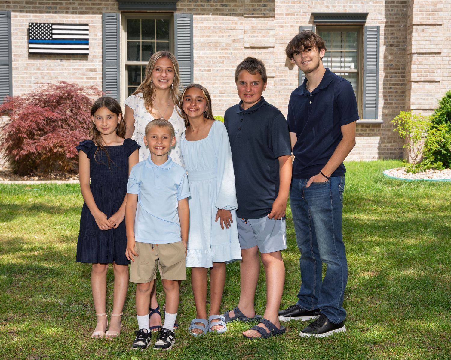 Six children of various ages pose together on a green lawn in front of a brick house on a sunny day.