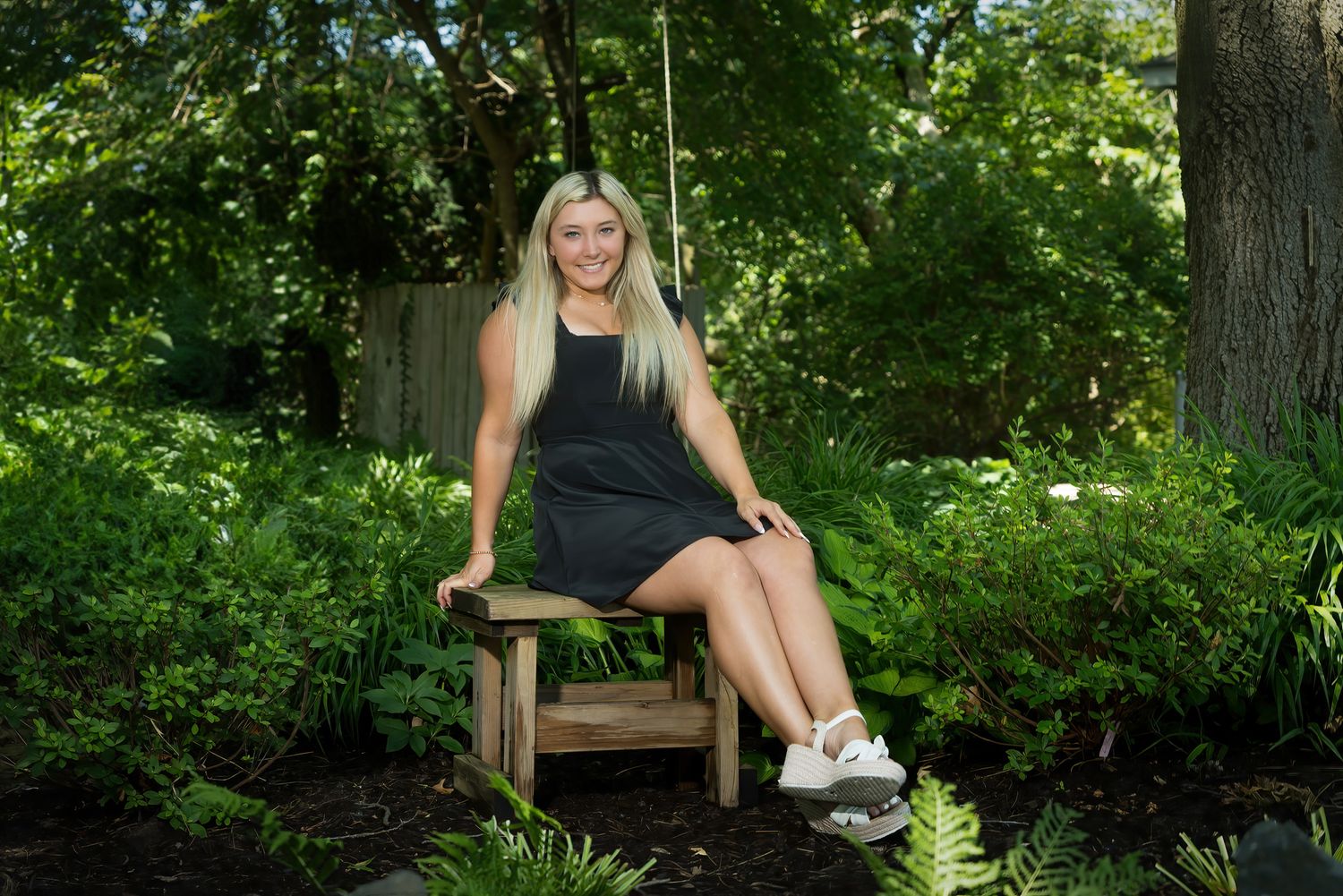 Cheerful blonde young woman sits on wooden garden bench surrounded by lush green foliage.