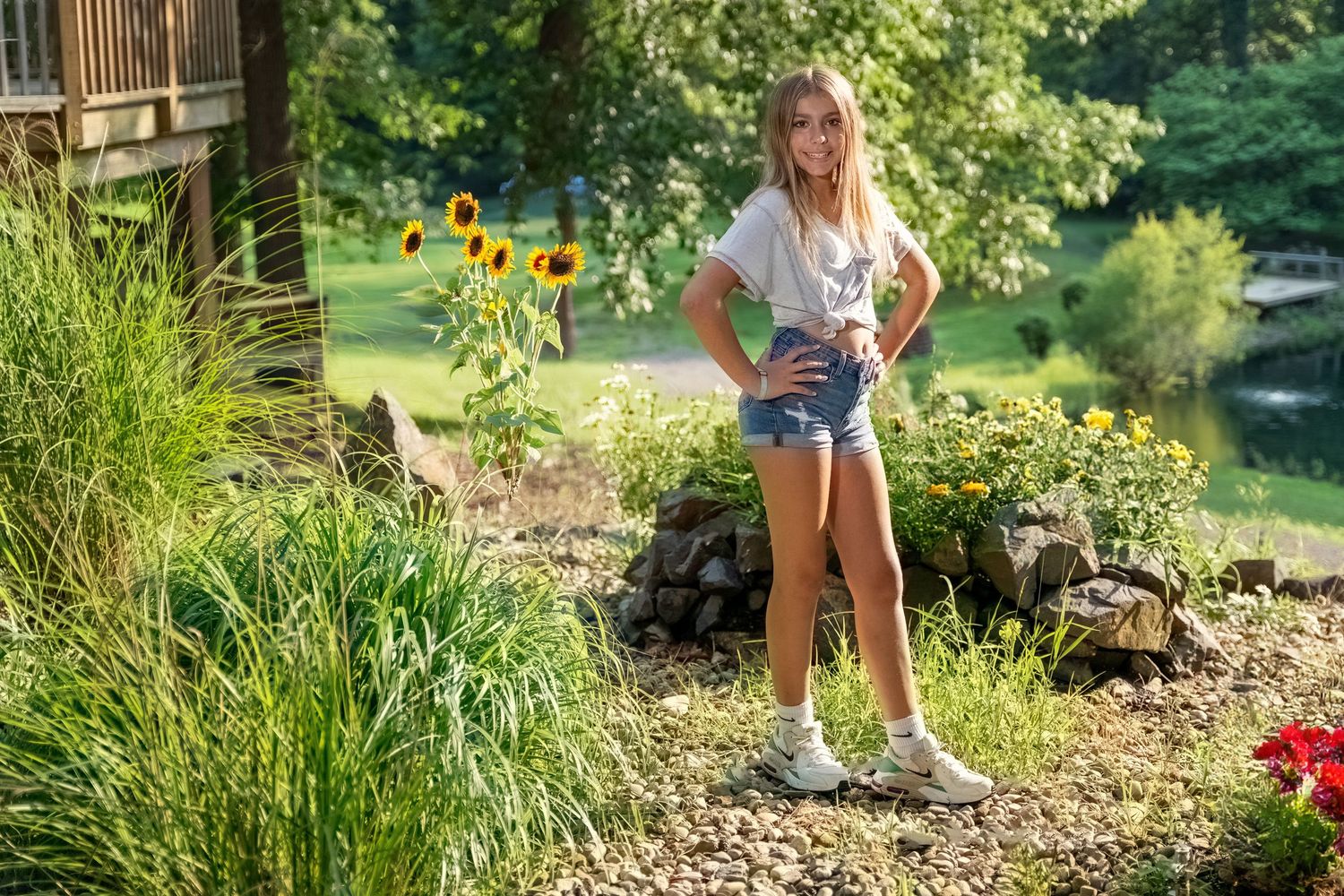 Young blonde girl in white tee and denim shorts stands among sunflowers in sunny garden setting.