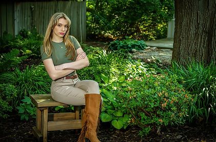 Young woman in olive top and khaki pants seated outdoors in lush green garden.