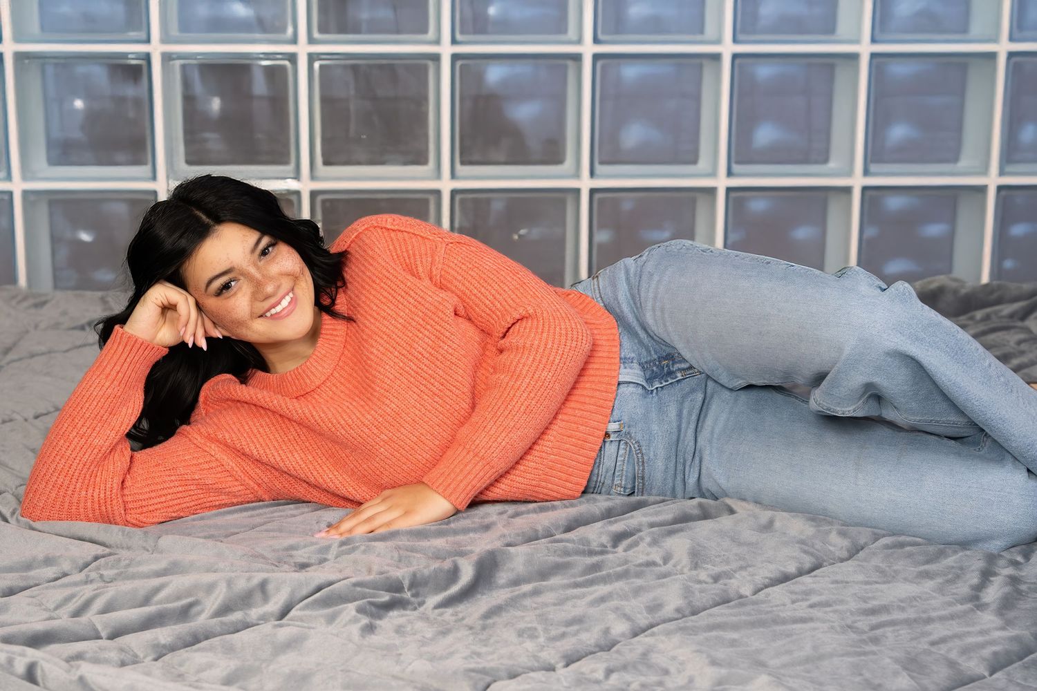 Young woman in an orange sweater and jeans smiling while lying on a gray surface.