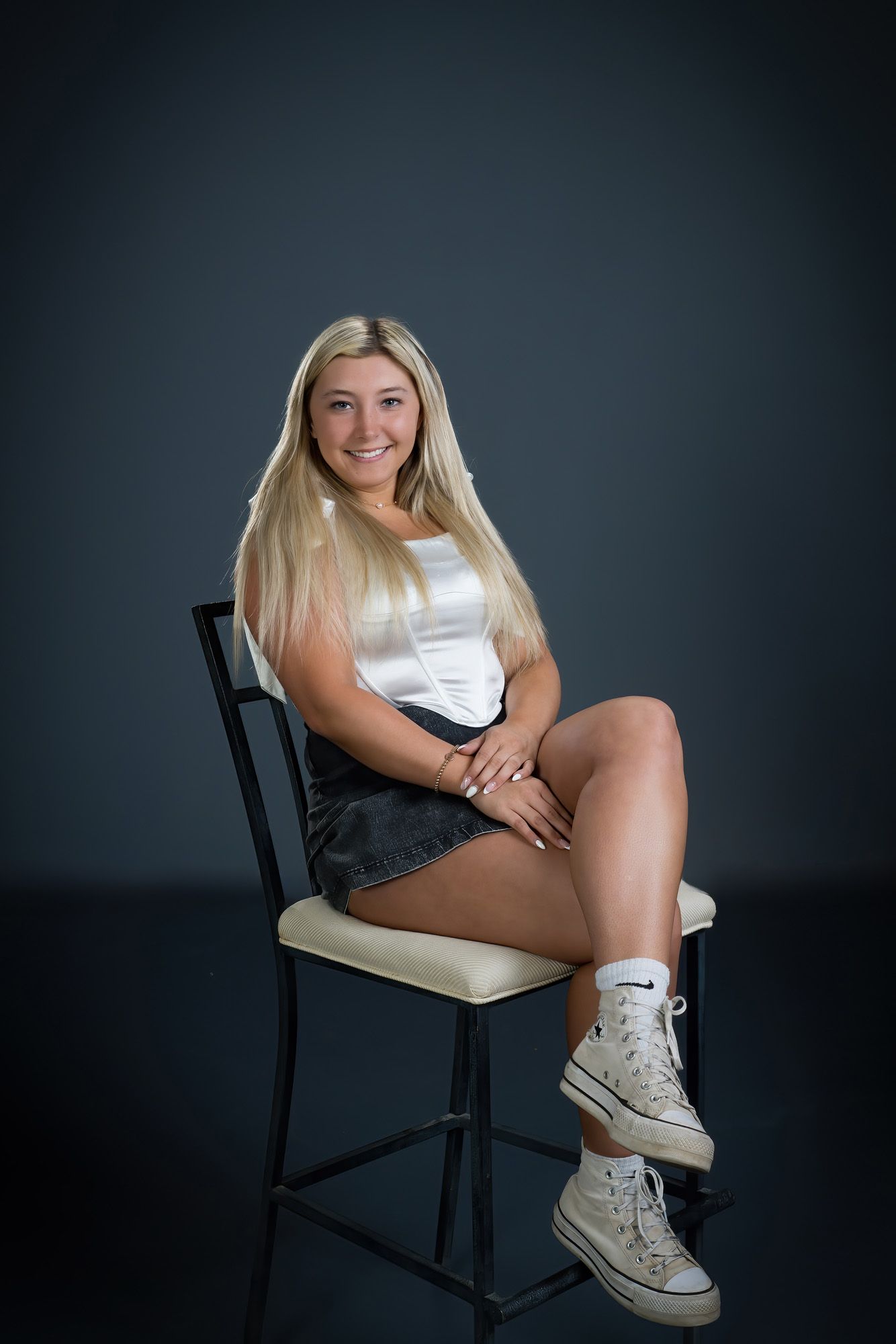 Smiling blonde teen in white top and denim skirt sitting on chair for senior portrait photo.