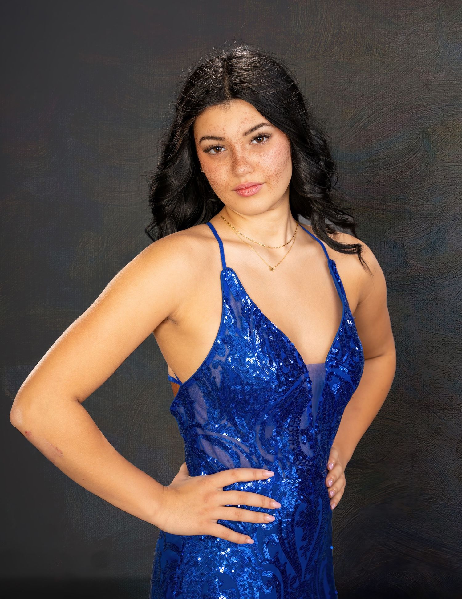 Young woman in a royal blue sequin v-neck formal gown posing confidently against a dark studio backdrop.