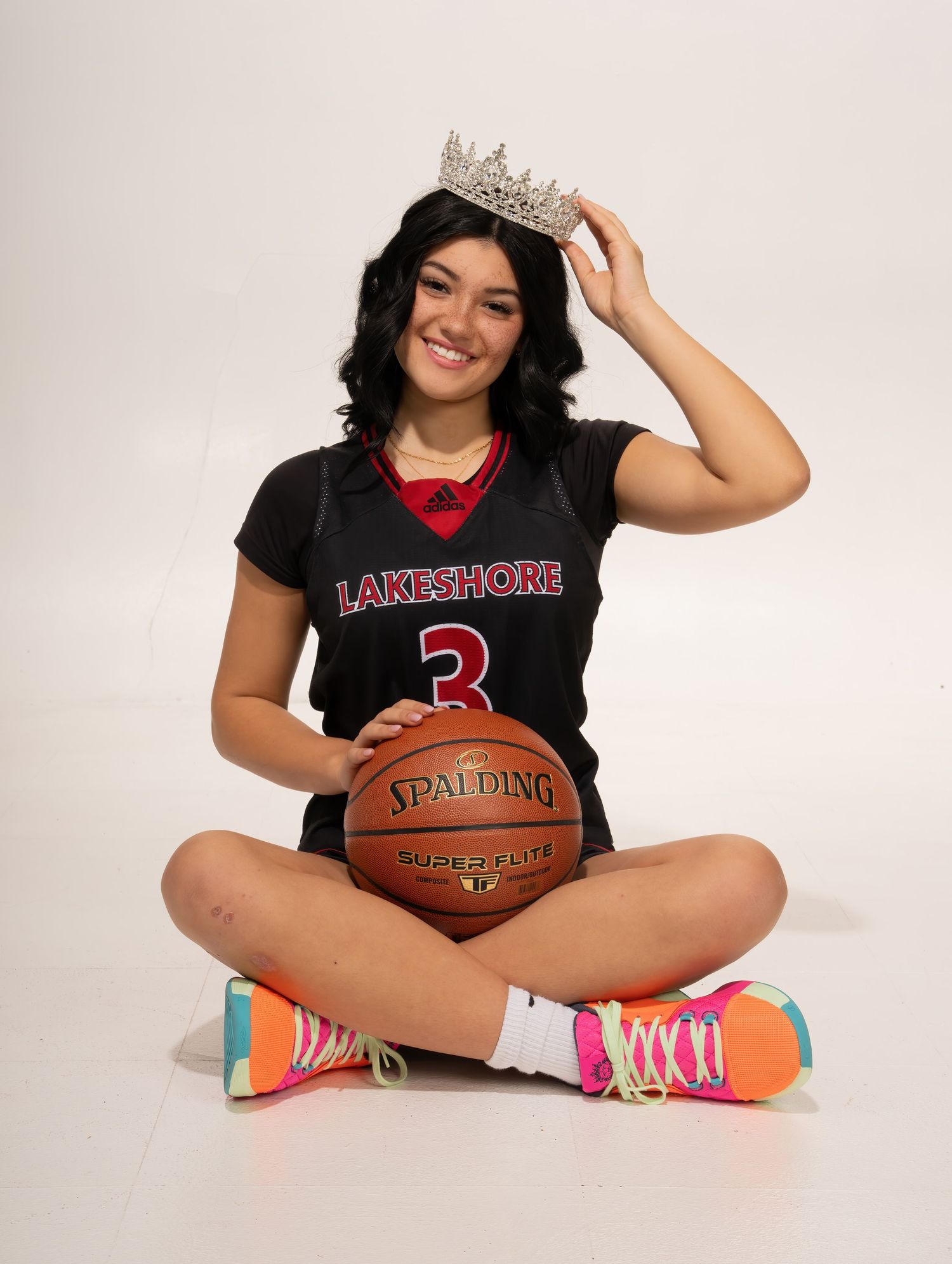 Athlete in Lakeshore jersey holding basketball wearing tiara, seated on floor.