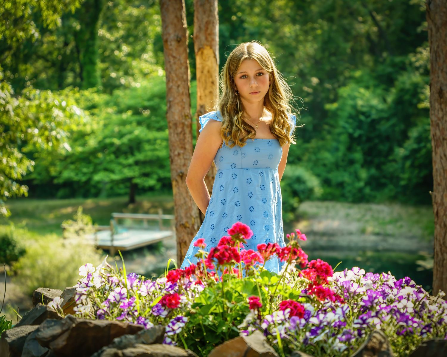 Young woman in blue sundress standing behind colorful red and purple flower garden near a serene pond.