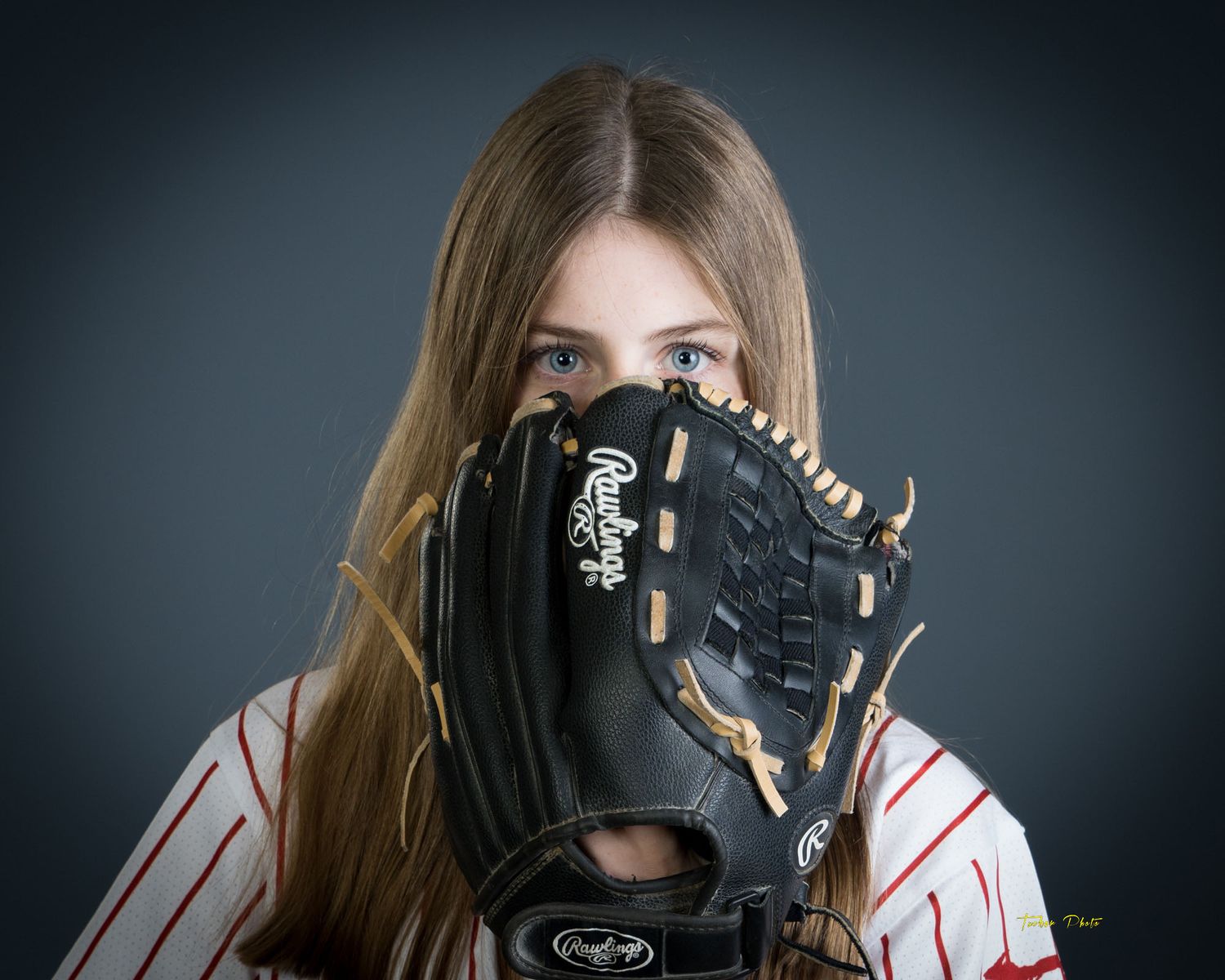 Blue-eyed girl in baseball uniform holding a black Rawlings glove up to her face on gray background.
