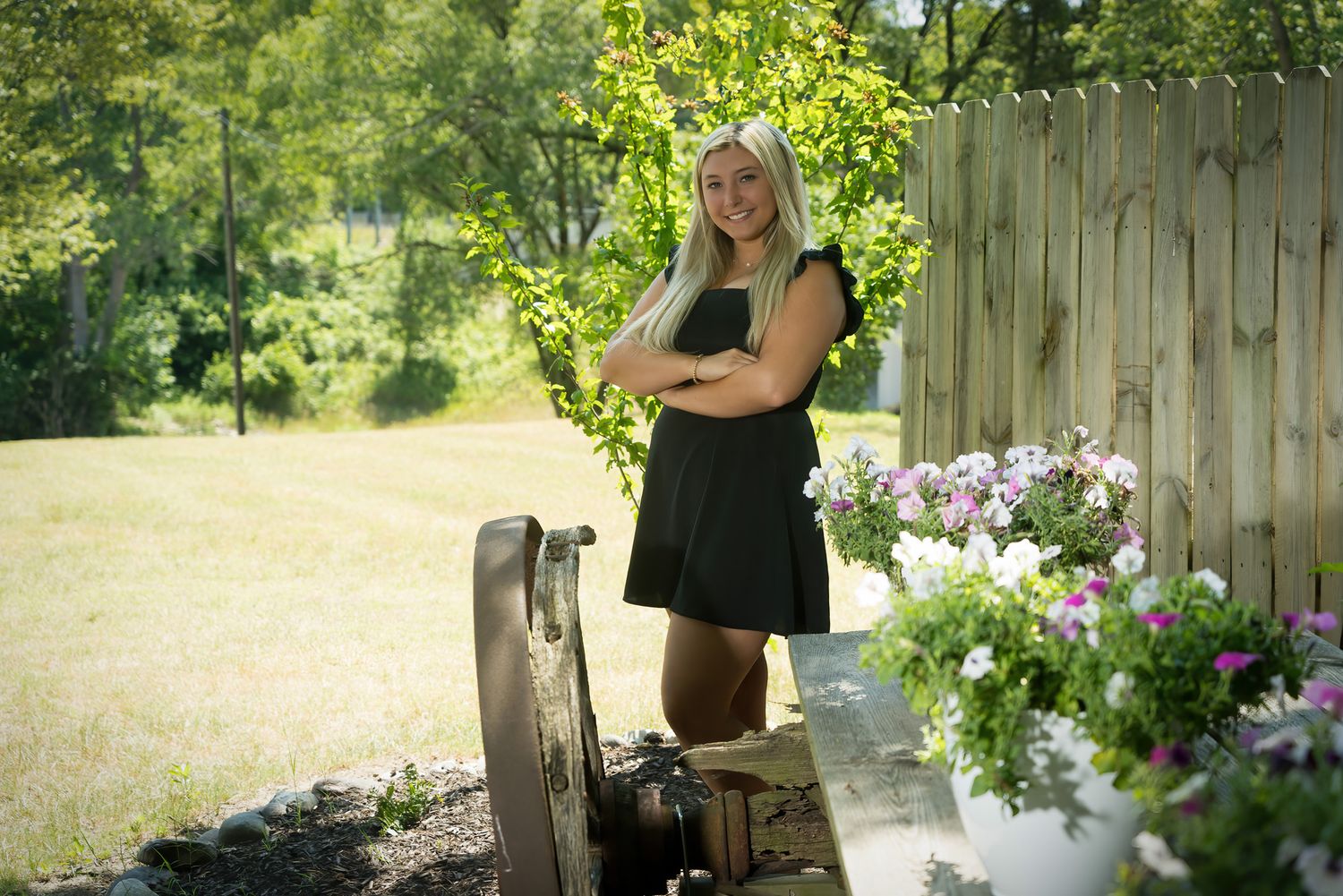 Smiling blonde girl in black dress poses outdoors near garden flowers and wooden fence.