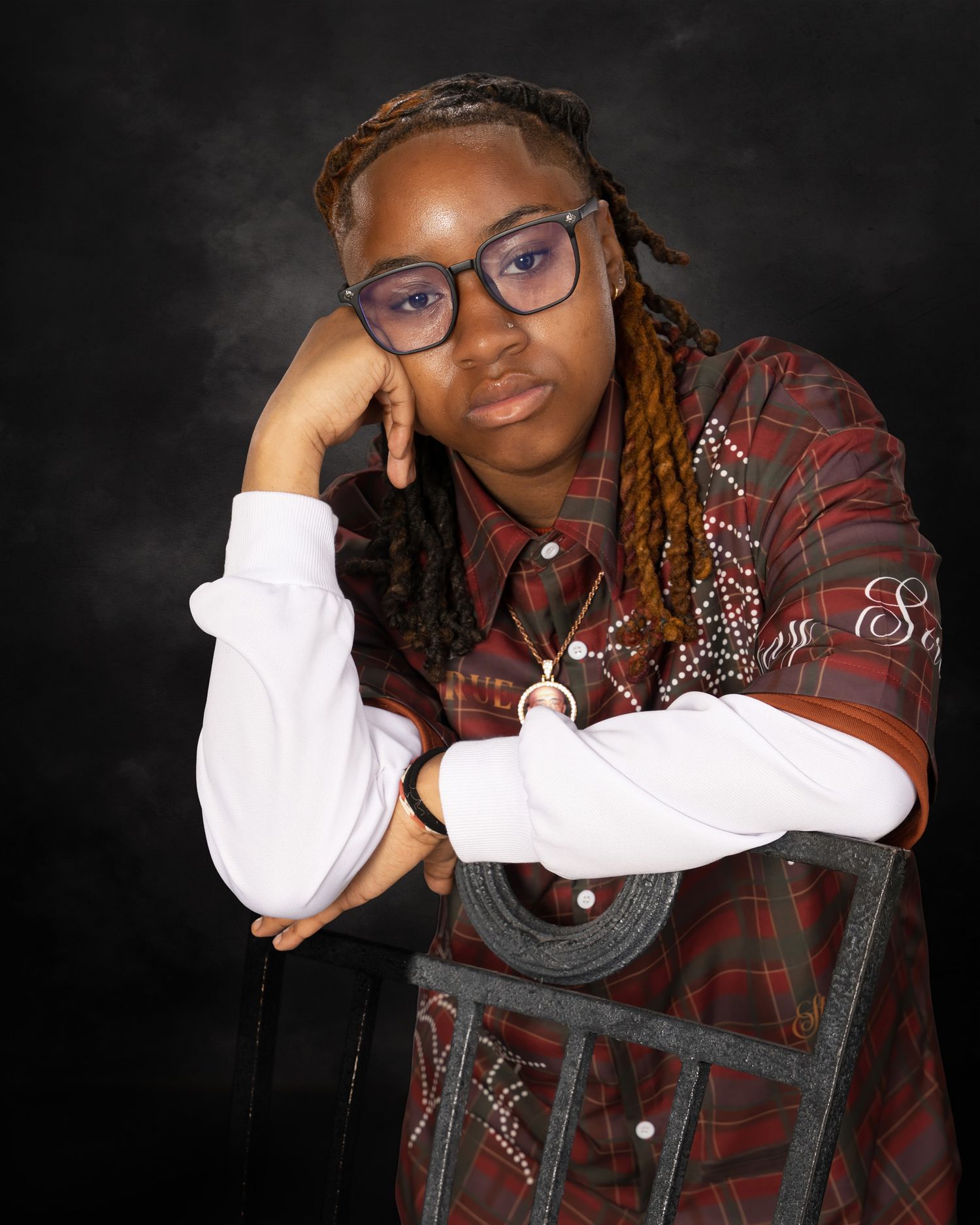 Young person with braids and glasses poses thoughtfully on vintage chair against dark studio background.