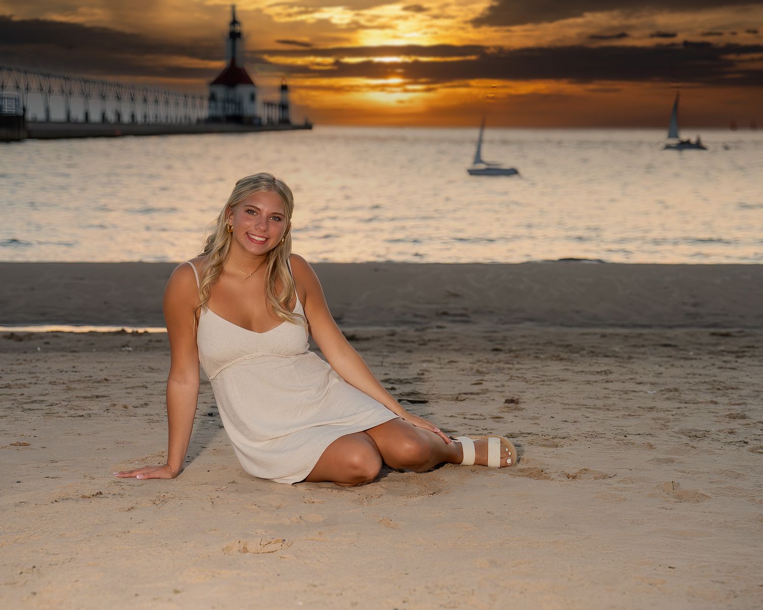 Blonde teenage girl in white dress sitting on sandy beach at dramatic sunset near pier.