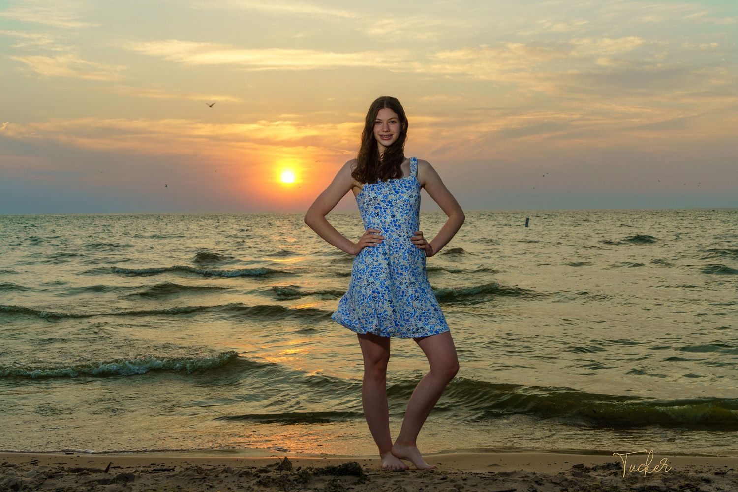 Teen girl in blue floral dress posing barefoot on sandy beach at colorful sunset.