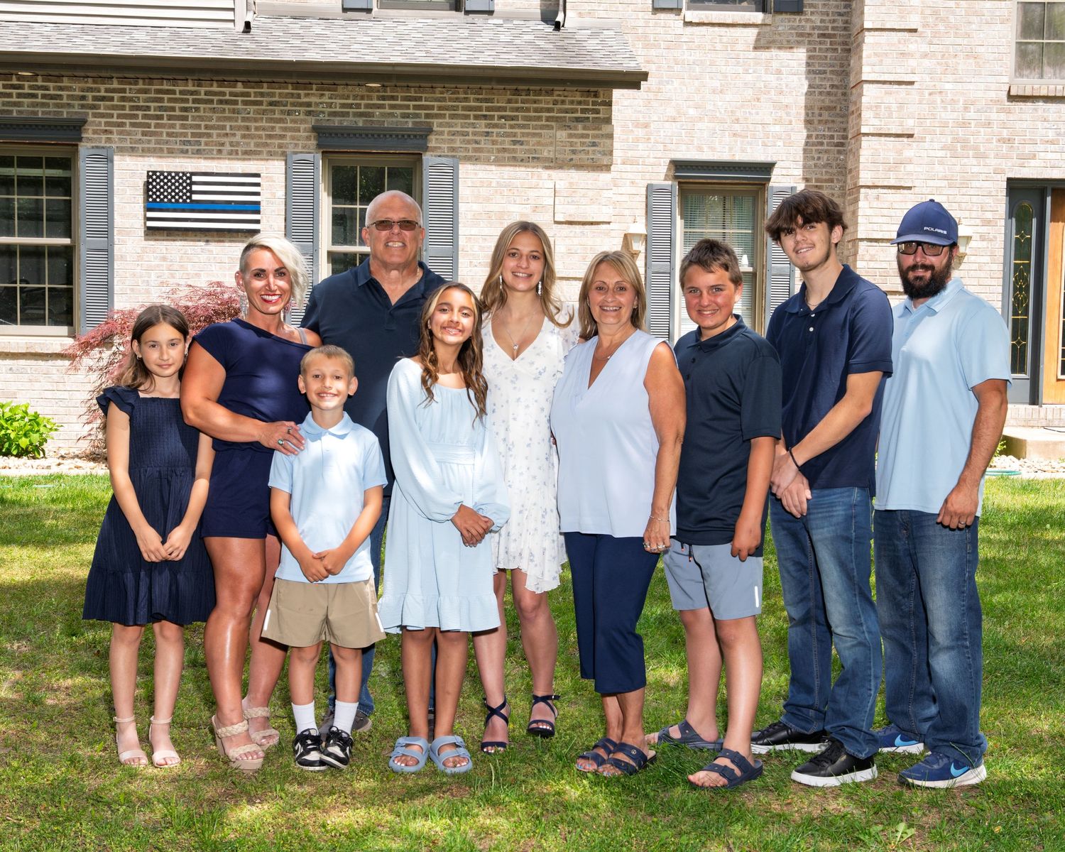 A large multigenerational family gathers for a group portrait on a green lawn outside a brick home.