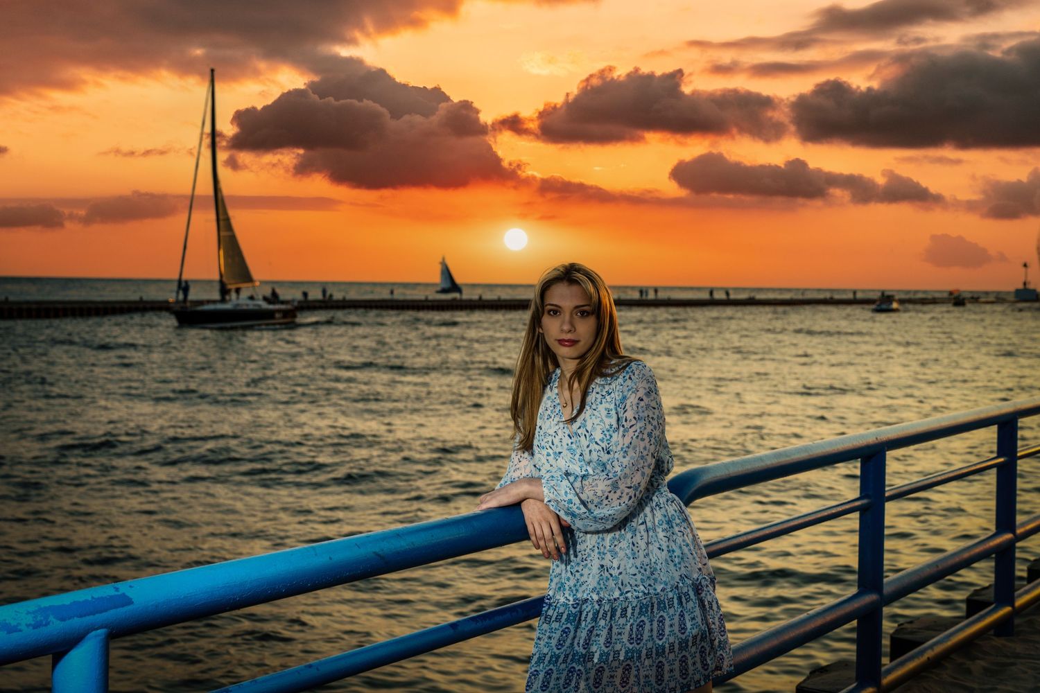 Young woman in floral dress leans on blue railing during stunning golden sunset over the ocean.