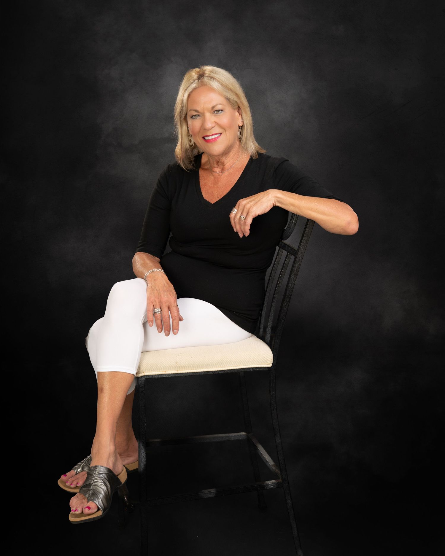 Elegant blonde woman in black top and white pants sitting gracefully on chair against dark background.