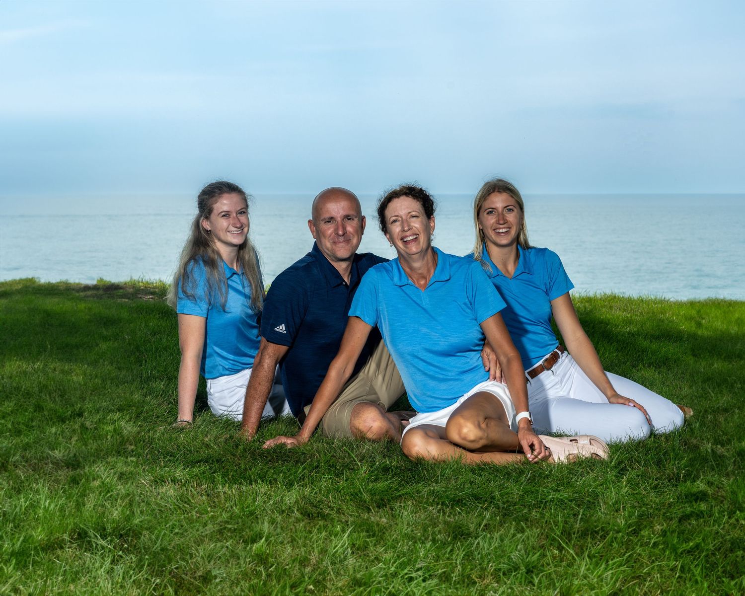 Family of four sitting together on green grass near ocean, wearing coordinated blue and white outfits.
