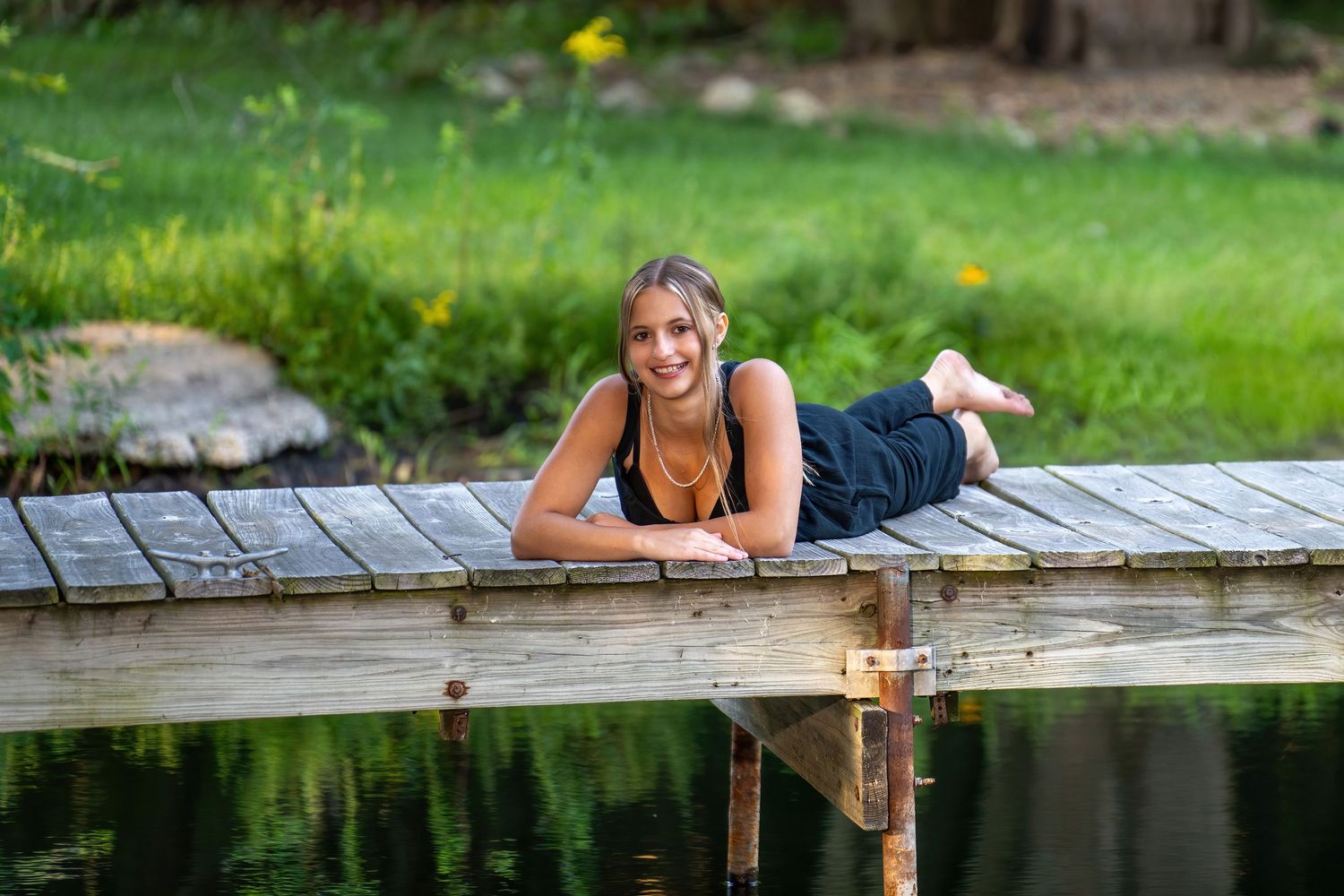 A smiling teenage girl in a navy jumpsuit relaxes on a wooden dock overlooking a serene reflective pond.