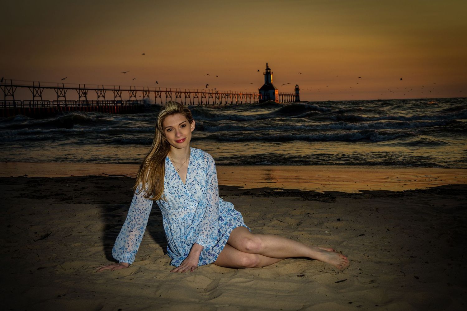Blonde teenage girl in floral dress sitting on beach at sunset near lighthouse pier.