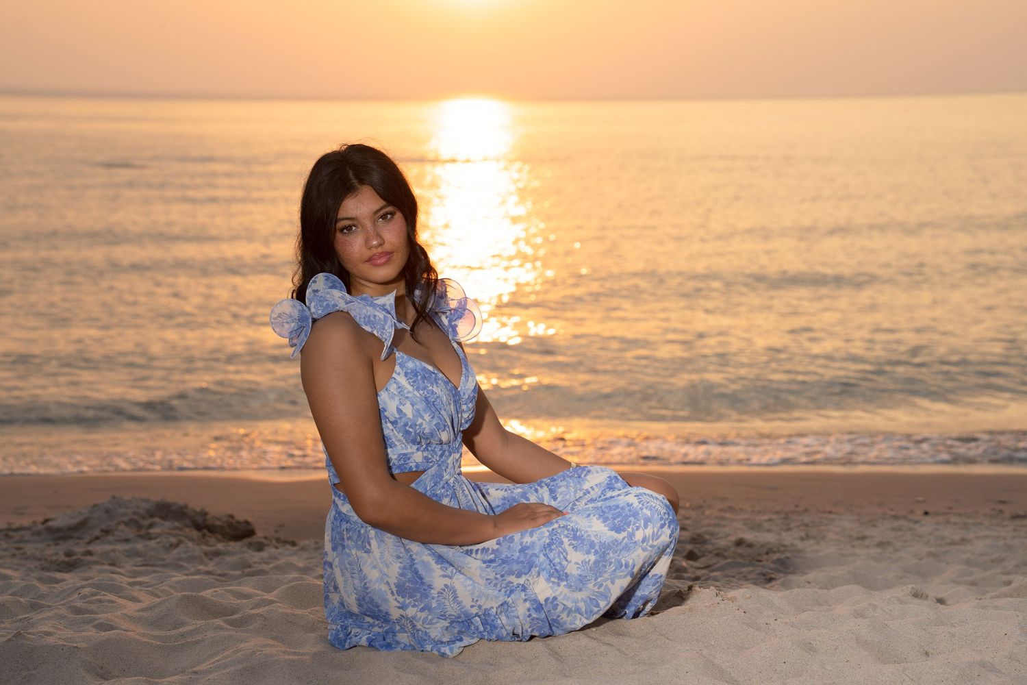 A young woman in a blue floral dress sits gracefully on a sandy beach during a golden sunset over calm waters.