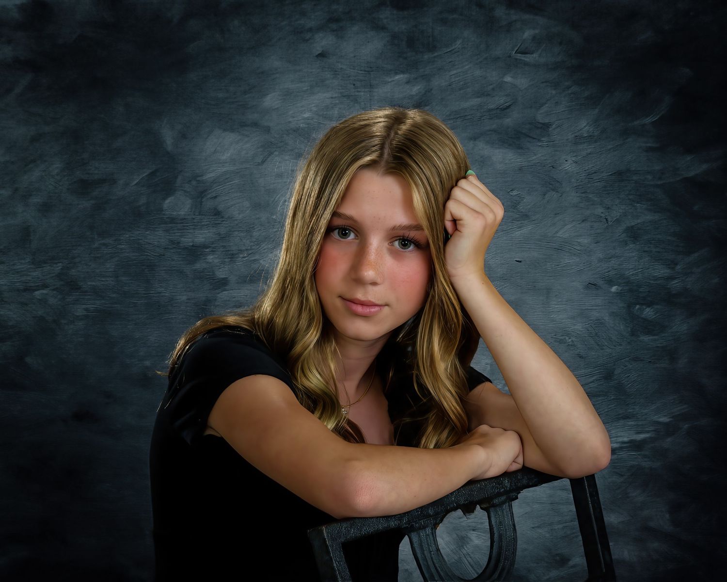 A young blonde girl in a black top rests her chin on her hand while posing against a textured blue-gray backdrop.