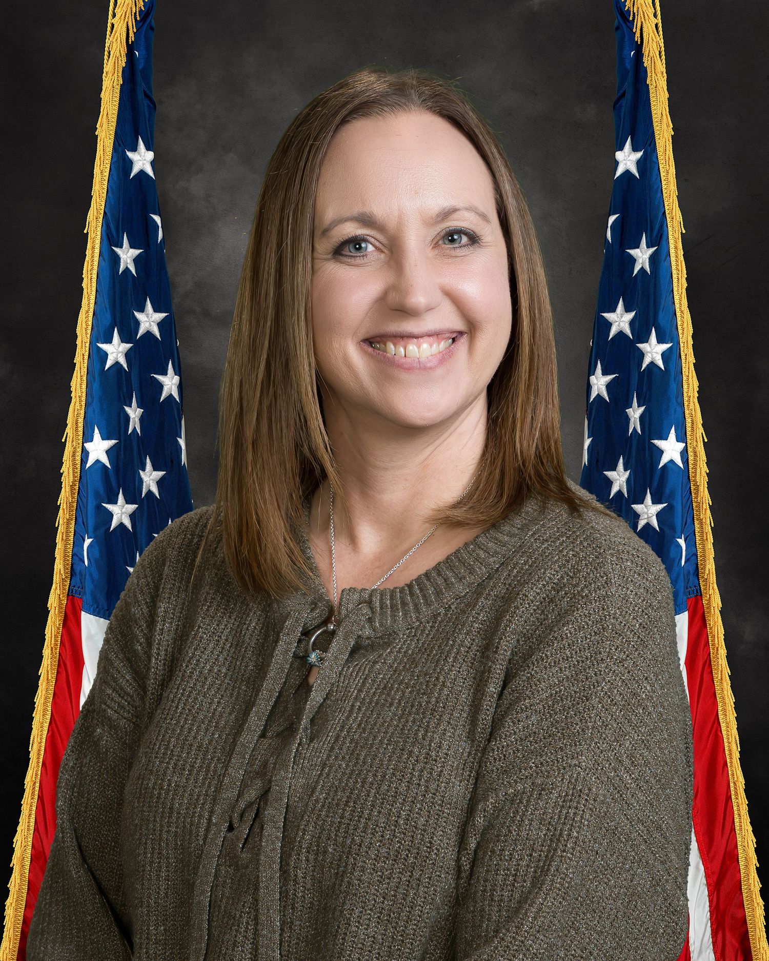 Professional portrait of a smiling woman with brown hair wearing a olive sweater before an American flag backdrop.