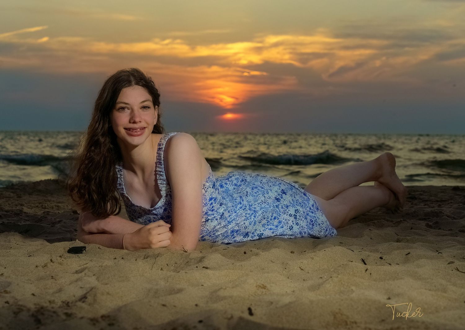 Young woman in blue floral dress lying on sandy beach with dramatic sunset over ocean waves.