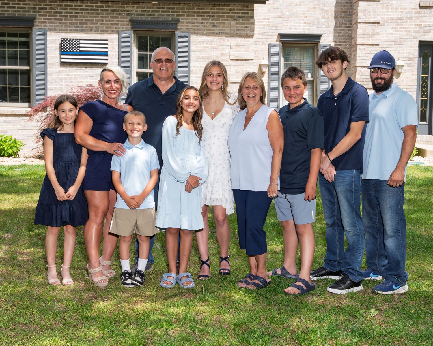 A large multigenerational family group poses together on a green lawn in front of a brick home.