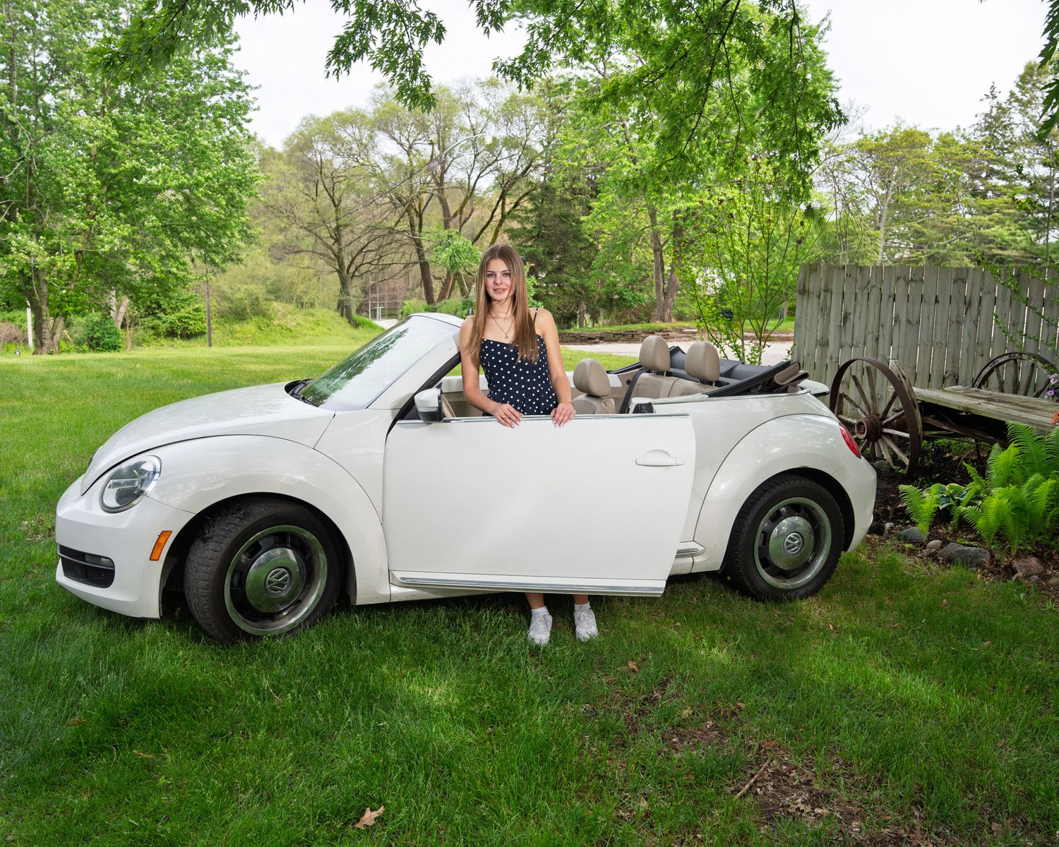 Young woman standing beside white VW Beetle convertible on green lawn outdoors.
