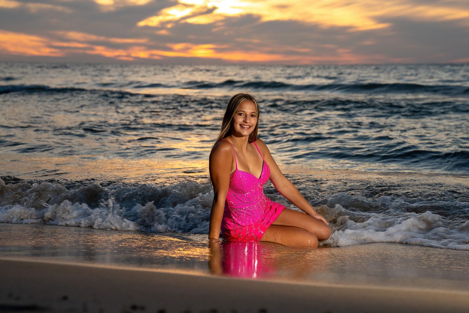 Smiling girl in pink dress sitting on sandy beach shore with waves and golden sunset.