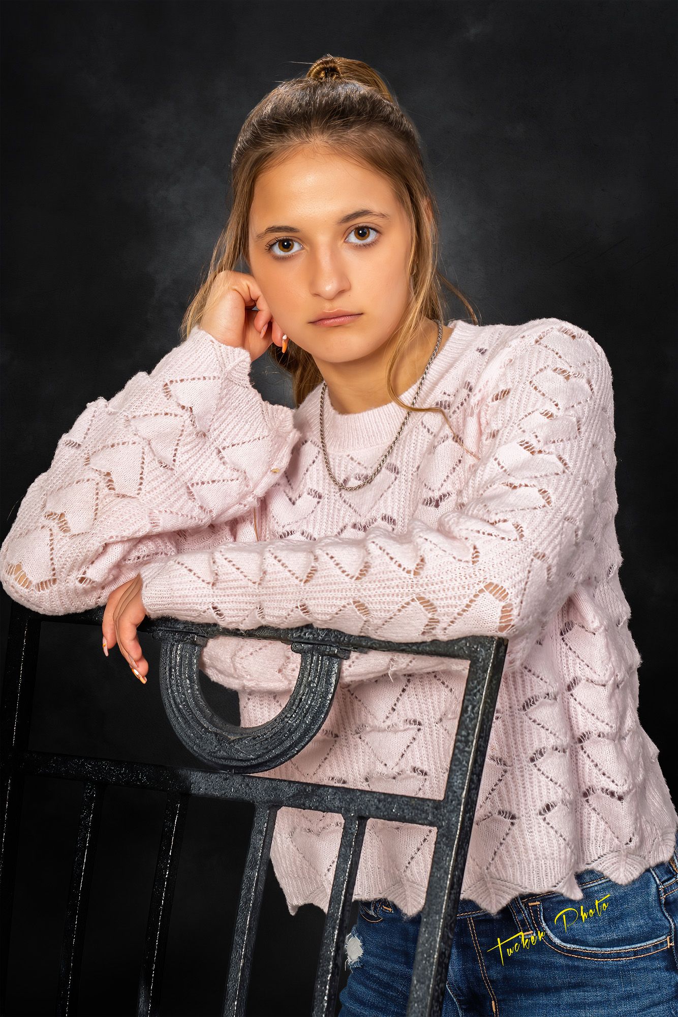 Teen girl in pink knit sweater leans thoughtfully on vintage black chair against dark studio backdrop.