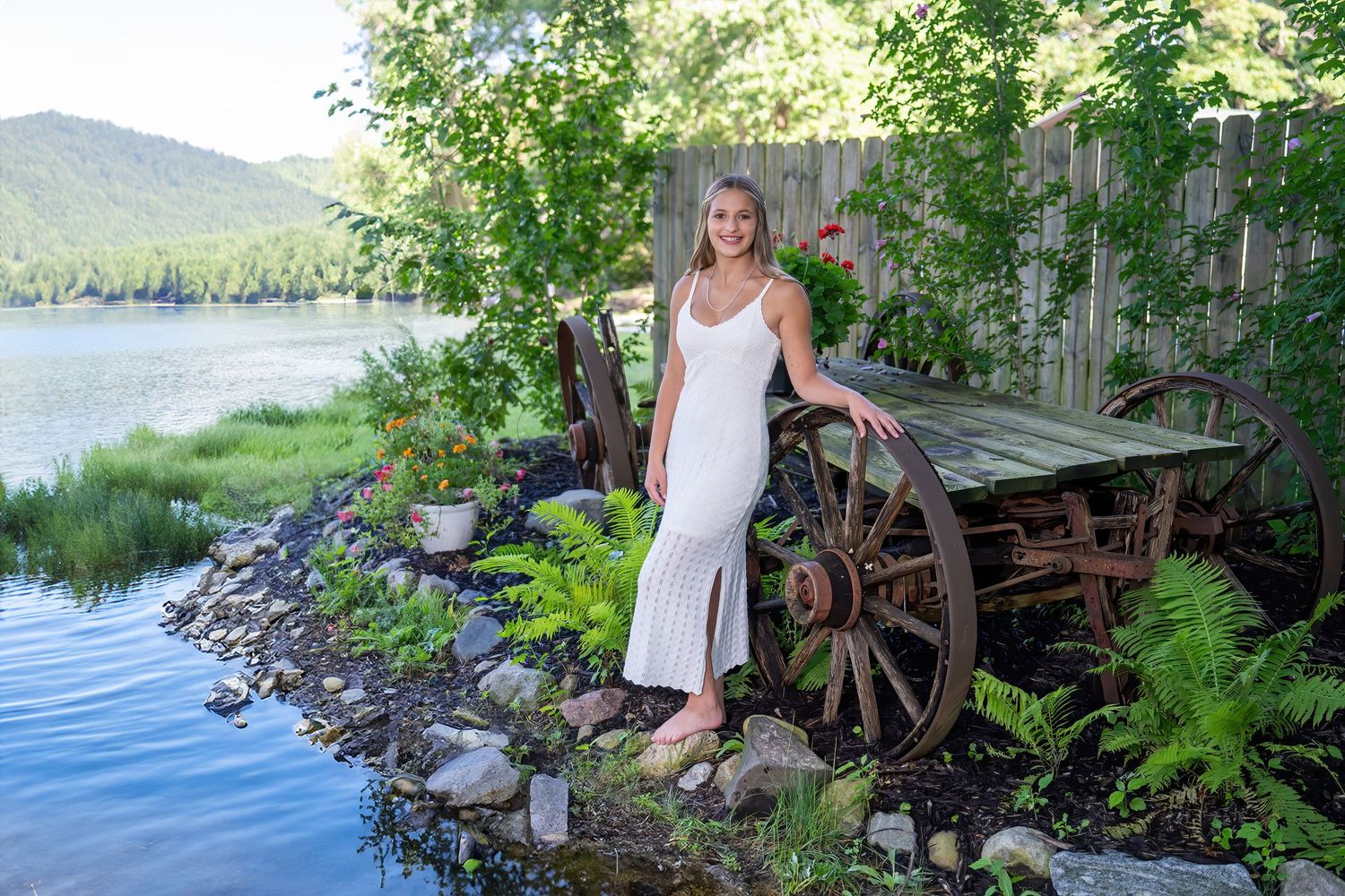 Young woman in white dress posing by rustic wooden wagon wheel near scenic lakeside garden.