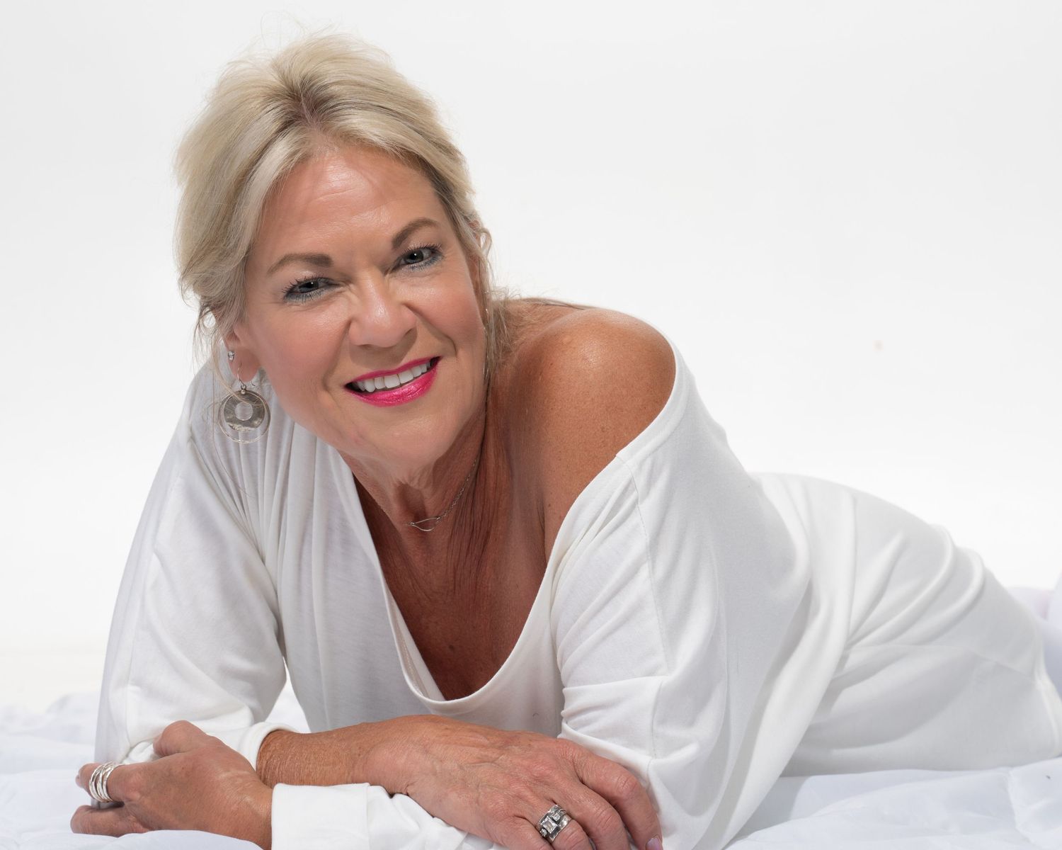 Smiling blonde woman in white off-shoulder top lying down on white background.
