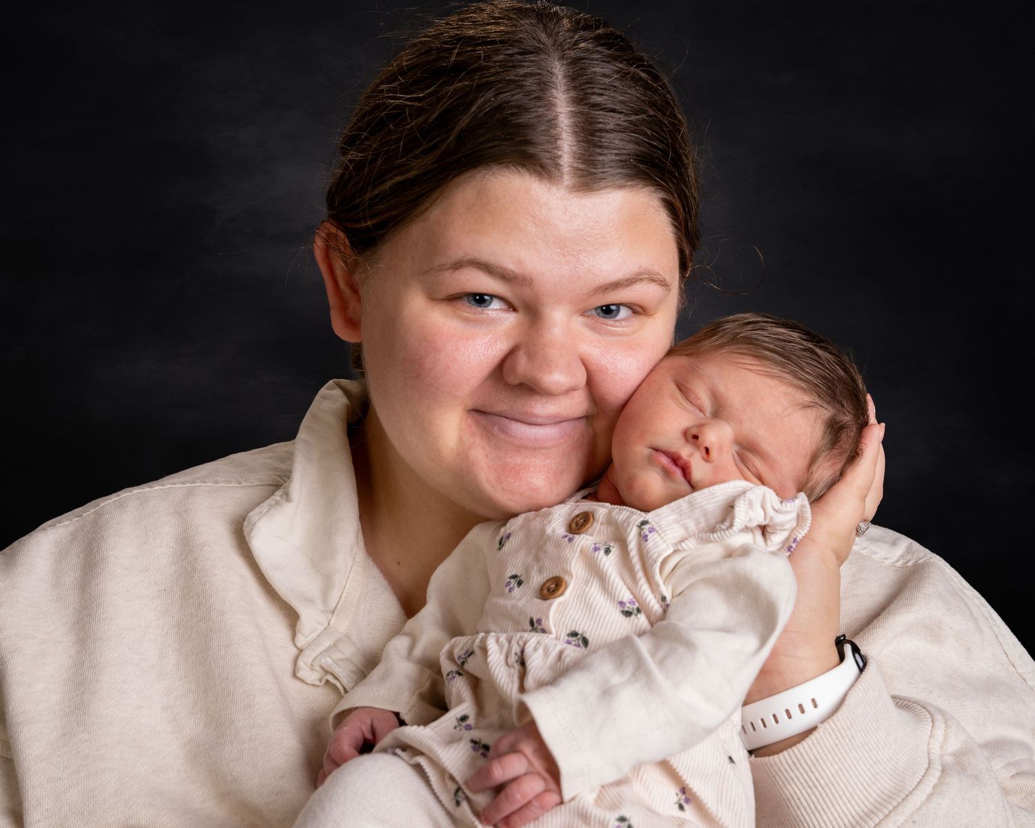 Smiling mother holding sleeping newborn baby against dark studio background.