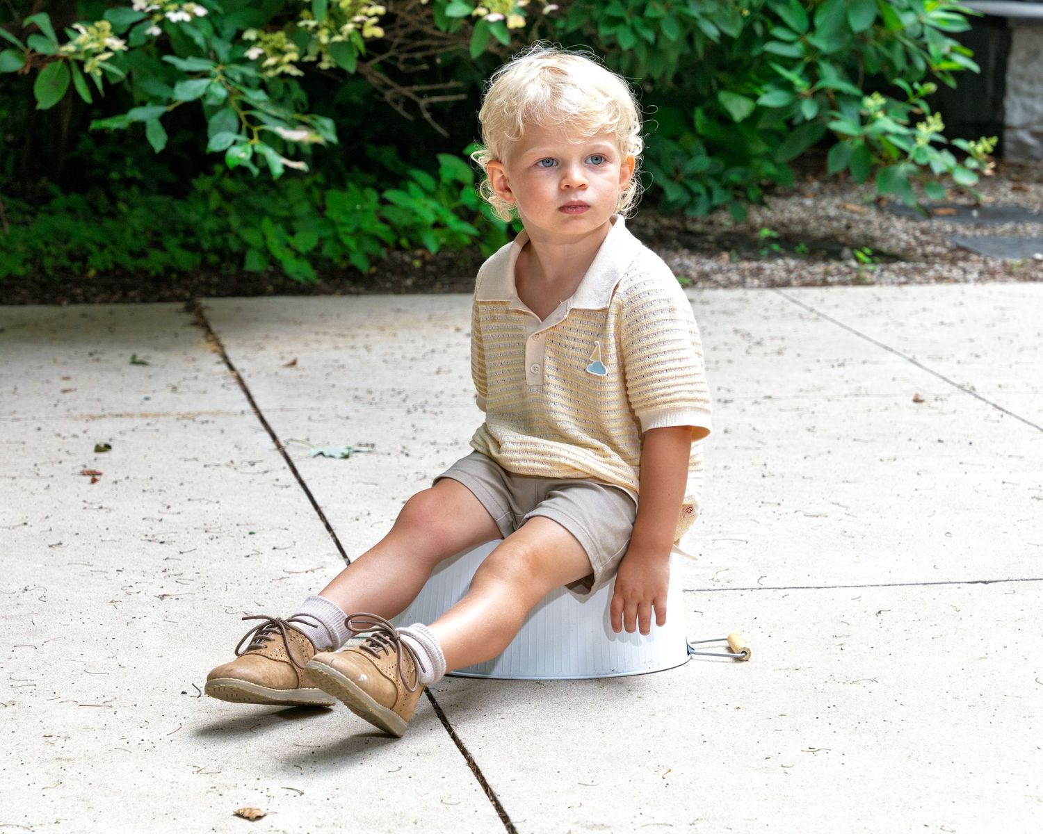 A cute blonde toddler boy in a beige polo shirt and shorts sits on a patio outdoors.
